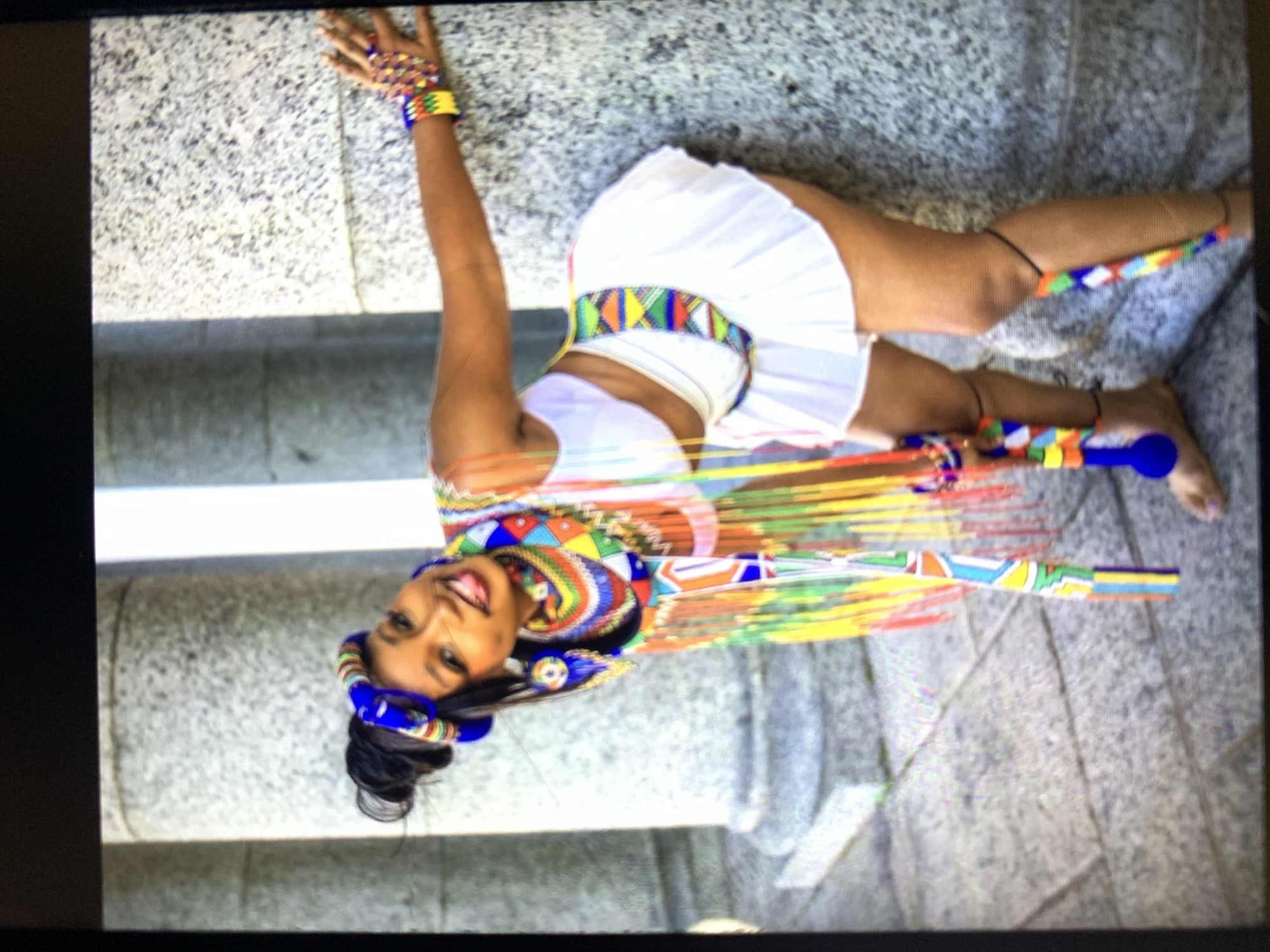 A woman is depicted lying on a marble or granite surface in an urban setting. She is adorned in traditional attire consisting of a white top, a white skirt, and colorful beaded accessories around her head, neck, waist, wrists, and ankles. A decorated baton or stick is placed on the surface near her feet. She is smiling toward the camera. The background is a blurred urban landscape. The location is specified as "New Stadium, Nigeria".