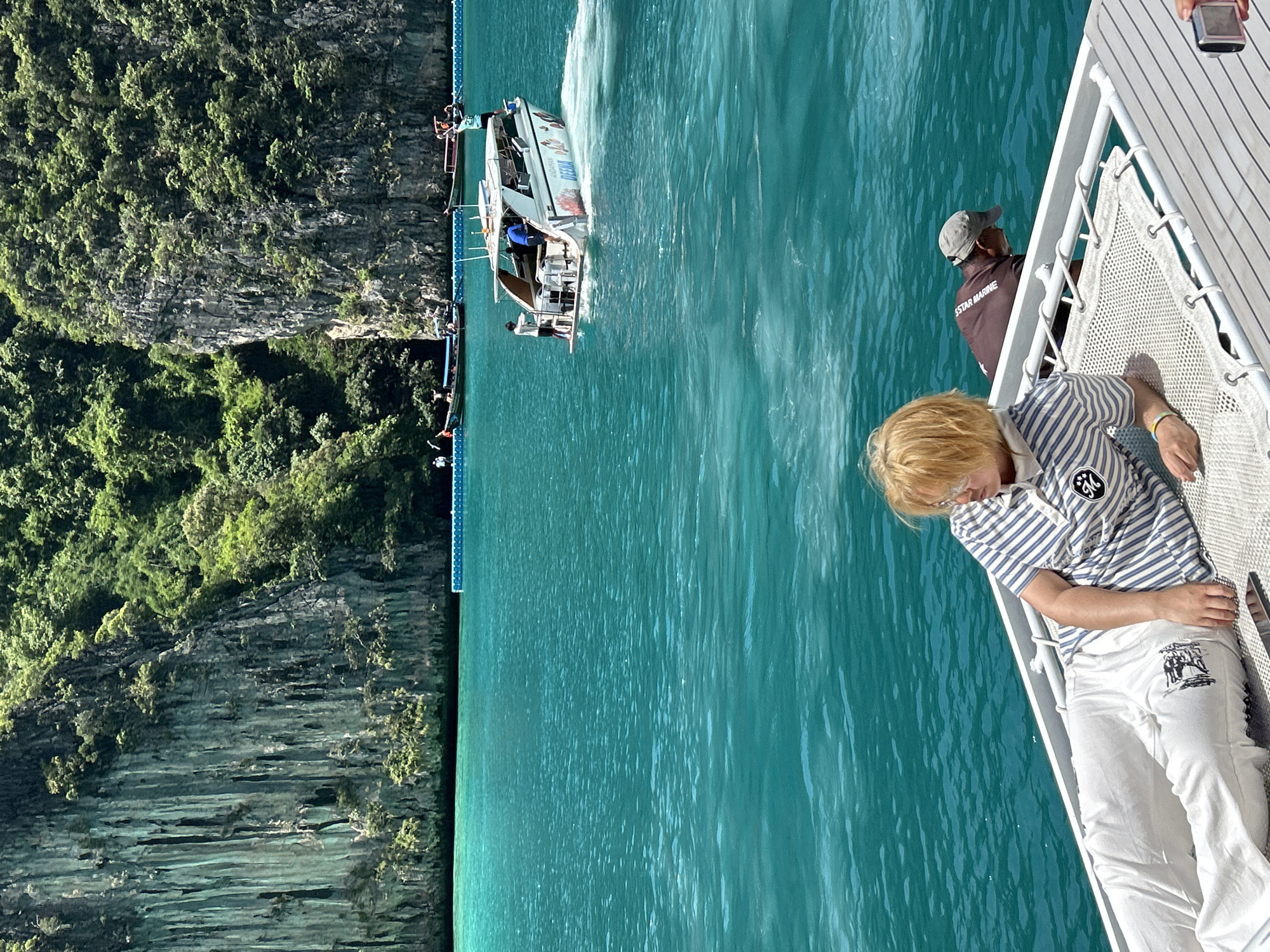 In Ao Nang, Thailand, a person with blonde hair wearing a striped polo shirt and white pants reclines on a white net hammock, facing the sea. The water is a vibrant turquoise, and in the background, a white speedboat with several people on board moves through the water, leaving a wake. Beyond the speedboat, lush green cliffs rise dramatically from the sea, covered in dense vegetation. Several longtail boats are also visible near the shore, and a blue floating barrier stretches across the water. Another person wearing a brown shirt and a grey cap is seated on the boat in the foreground, behind the person on the hammock. The overall scene depicts a sunny day with clear skies and calm seas in a tropical coastal environment.