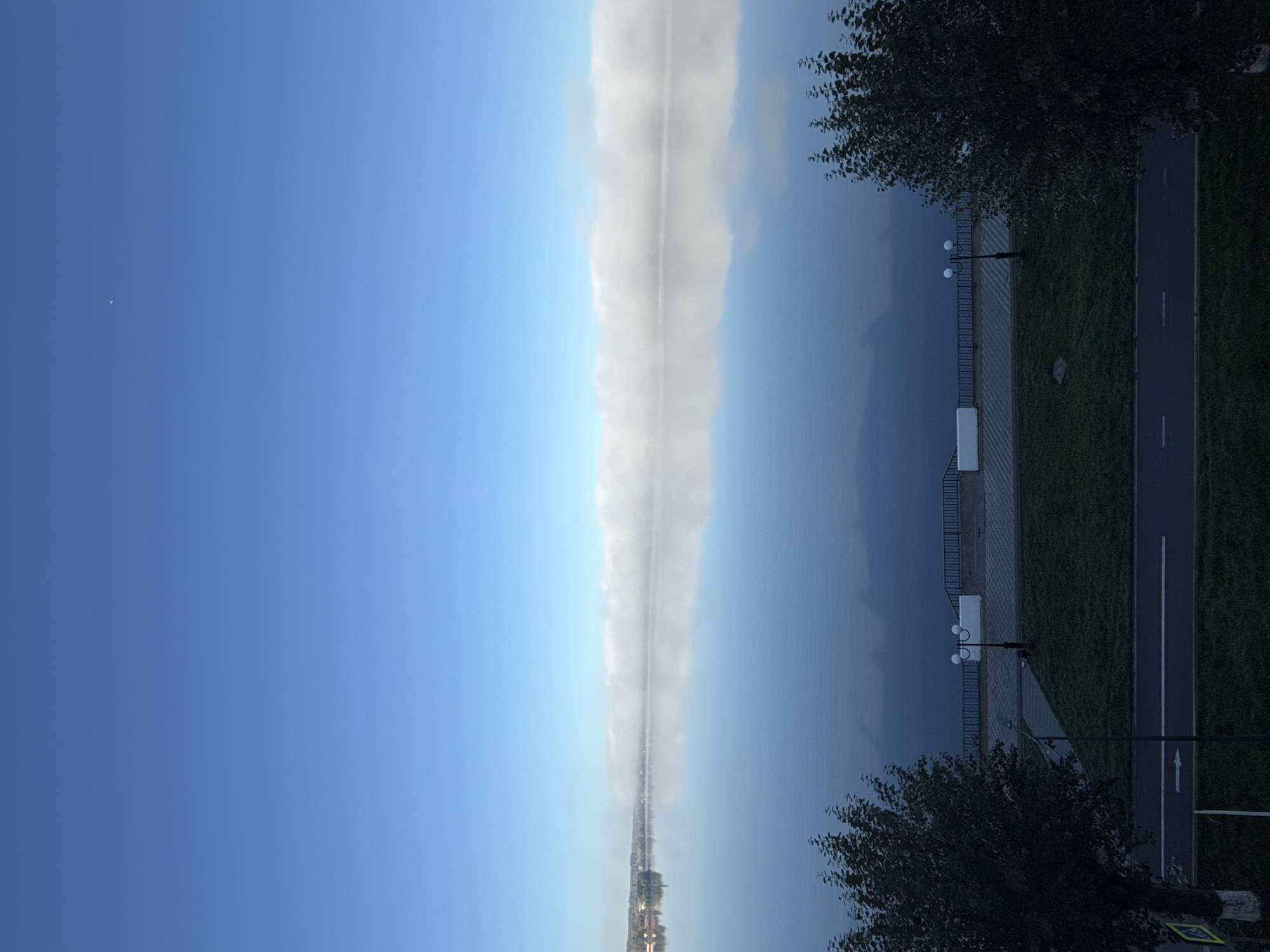 This image depicts a serene, early morning view of the Votkinsk Reservoir in Votkinsk, Russia. The sky is a clear, pale blue, with a faint hint of a star visible in the upper right quadrant. A band of low-lying white mist stretches horizontally across the middle distance, obscuring the far shore and creating a reflective, mirror-like surface on the calm water. On the left side of the frame, a distant town with red-roofed buildings is partially visible through the mist.
In the foreground, a paved walkway with decorative lampposts runs parallel to the water's edge. This walkway is bordered by a neatly mowed green lawn. Two trees, one on the left and one on the right, frame the scene. A paved road with white lane markings, presumably a bike path or pedestrian walkway, is visible at the bottom of the image, with a yellow traffic sign on the far left. The overall atmosphere is tranquil and quiet, suggesting an undisturbed start to the day. There are no people or animals visible in the image.