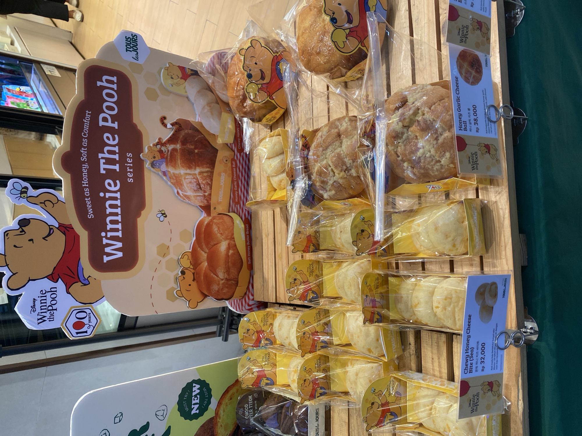 This image shows a display of baked goods, likely from a bakery, with a Winnie the Pooh theme. The products are arranged on wooden shelves and are individually wrapped in clear plastic. Many of the packages feature illustrations of Winnie the Pooh and friends.
The prominent display sign on the left reads "Disney Winnie The Pooh series," with smaller text "Sweet as Honey, Soft as Comfort." It also mentions "100 years," suggesting a celebration related to Winnie the Pooh's anniversary.
Several types of bread and pastries are visible. One sign indicates "Soft Honey Butter Bread." Other visible product names include "Honey Garlic Cheese Ball" priced at Rp 38,000, and "Chewy Honey Cheese Bite (3ea)" priced at Rp 32,000. The text on these signs includes both English and Korean.
The overall setting appears to be indoors, likely a retail environment. The lighting suggests it could be daytime. The presence of price tags in Indonesian Rupiah (Rp) and the text "North Jakarta, Indonesia" in the context clues confirm the location. There are no people clearly visible in the frame, focusing entirely on the food products and promotional materials. The scene is clean and organized, typical of a bakery display.
