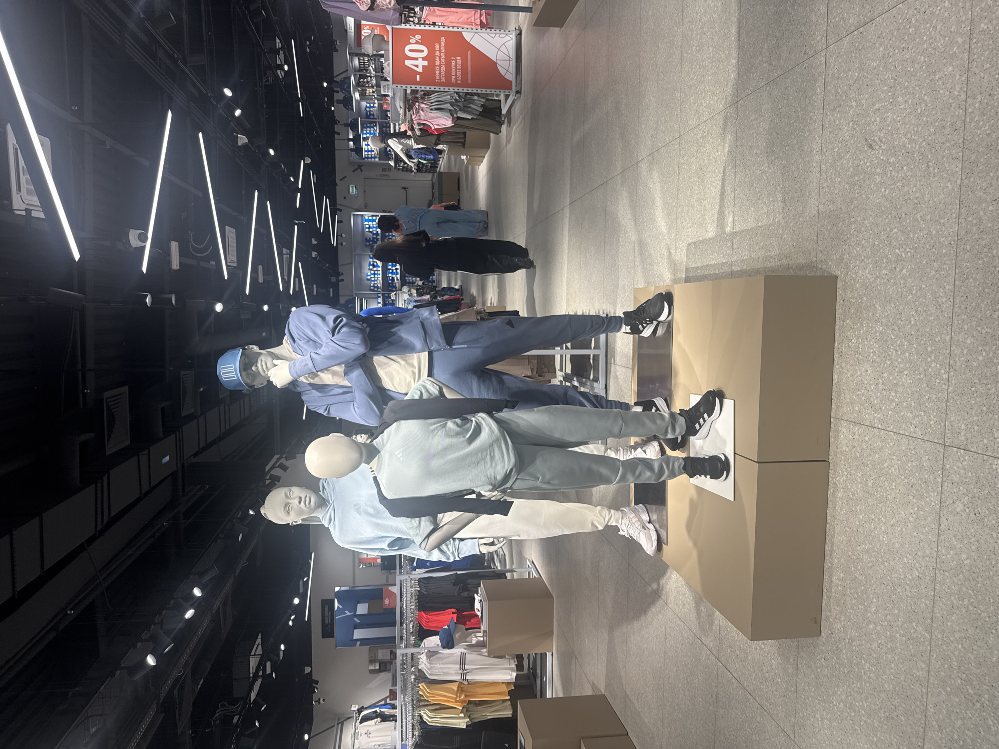 This media file depicts a retail store interior, likely a clothing store, with a focus on athletic wear. In the foreground, there are several mannequins dressed in casual sportswear. The mannequins are arranged in a way that suggests they are displaying outfits.
One mannequin is wearing a blue tracksuit and a blue cap. Next to it, another mannequin is dressed in a light blue hoodie and light-colored pants, paired with white sneakers. A third mannequin is wearing a light blue top and dark gray pants, also with black sneakers. The clothing displays are placed on neutral-colored pedestals.
In the background, racks of clothing are visible, with various t-shirts in different colors, including red, yellow, and orange. A prominent sign in the background features a "40%" discount offer, with text in what appears to be a Cyrillic-based alphabet, "2 НЕМЕСЕ ОДАН ДА КӨП ЗАТТАРДЫ САТЫП АЛҒАНДА ПРИ ПОКУПКЕ 2 И БОЛЕЕ ВЕЩЕЙ," which translates to "When buying 2 or more items." This indicates a sale or promotion.
The store appears to be well-lit with artificial lighting, including long strip lights on the ceiling and spotlights directed at the displays. The floor is tiled with a speckled gray pattern. The overall atmosphere suggests a modern and organized retail environment. The context provided indicates the location is Kokshetau, Kazakhstan. There are no discernible elements suggesting time of day or weather conditions, as the scene is indoors. The emotions conveyed are neutral, typical of a commercial setting.