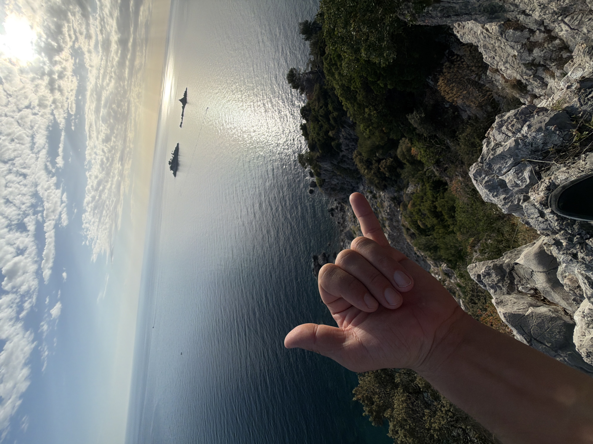 A wide-angle, eye-level shot captures a landscape view featuring a rocky terrain leading down to the sea in Petrovac centar, Montenegro. In the foreground, a right hand is making the "hang loose" gesture. The sea reflects sunlight creating a shimmering effect, with two small islands visible in the distance. The sky is partially covered with clouds with the sun shining through. The foreground contains rocky terrain with greenery along the cliff. A small portion of a shoe is visible at the top right corner of the image.