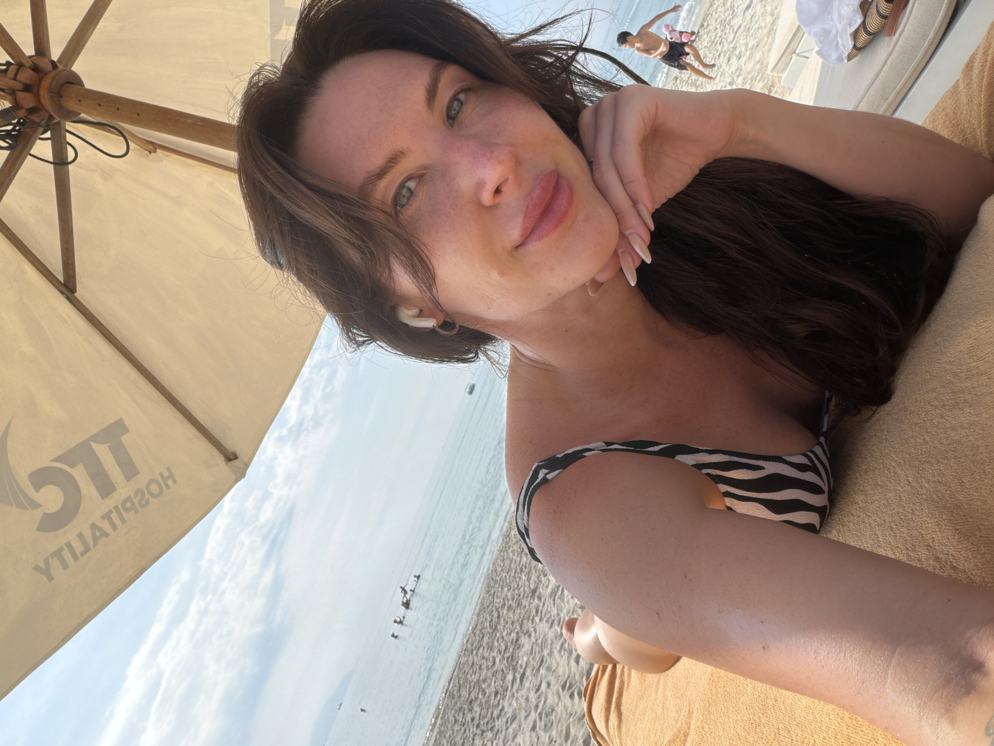 A woman is lying on a beach towel under a beach umbrella at a beach in Ninh Hoa Ward, Dong Hoi City, Vietnam. She is wearing a black and white striped bikini top and has her right hand resting on her chin. She is looking towards the camera with a slight smile. In the background, the ocean is visible with several people swimming and a child running on the sand. The beach umbrella has the words "TLC HOSPITALITY" printed on it. The scene is bright and sunny, with a clear blue sky.