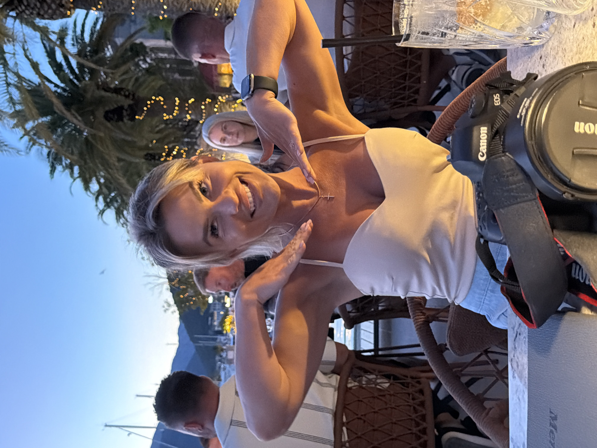 A blonde woman with fair skin and bright blue eyes is seated at an outdoor table at dusk in Tivat, Montenegro. She is smiling and looking at the camera, her left hand near her neck and her right hand resting on her chest. She is wearing a light-colored tank top with thin straps and a cross necklace. Her arms are visible, along with a black Apple Watch on her right wrist. In front of her, on the table, is a black Canon EOS camera with a strap.
In the background, other people are seated at tables. To the left, a man wearing a striped shirt is partially visible, and further left, a boat mast is seen against the twilight sky. To the right of the woman, a blonde woman with light skin and blue eyes is seated, looking ahead. In the far right of the background, another man in a white shirt is visible.
The setting appears to be a restaurant or bar with palm trees adorned with fairy lights in the background. The sky is a deep blue, indicating it is evening. The table has a marble-like surface. A glass with ice and a drink is on the right side of the table. The chairs are made of wicker. The overall atmosphere is social and relaxed.