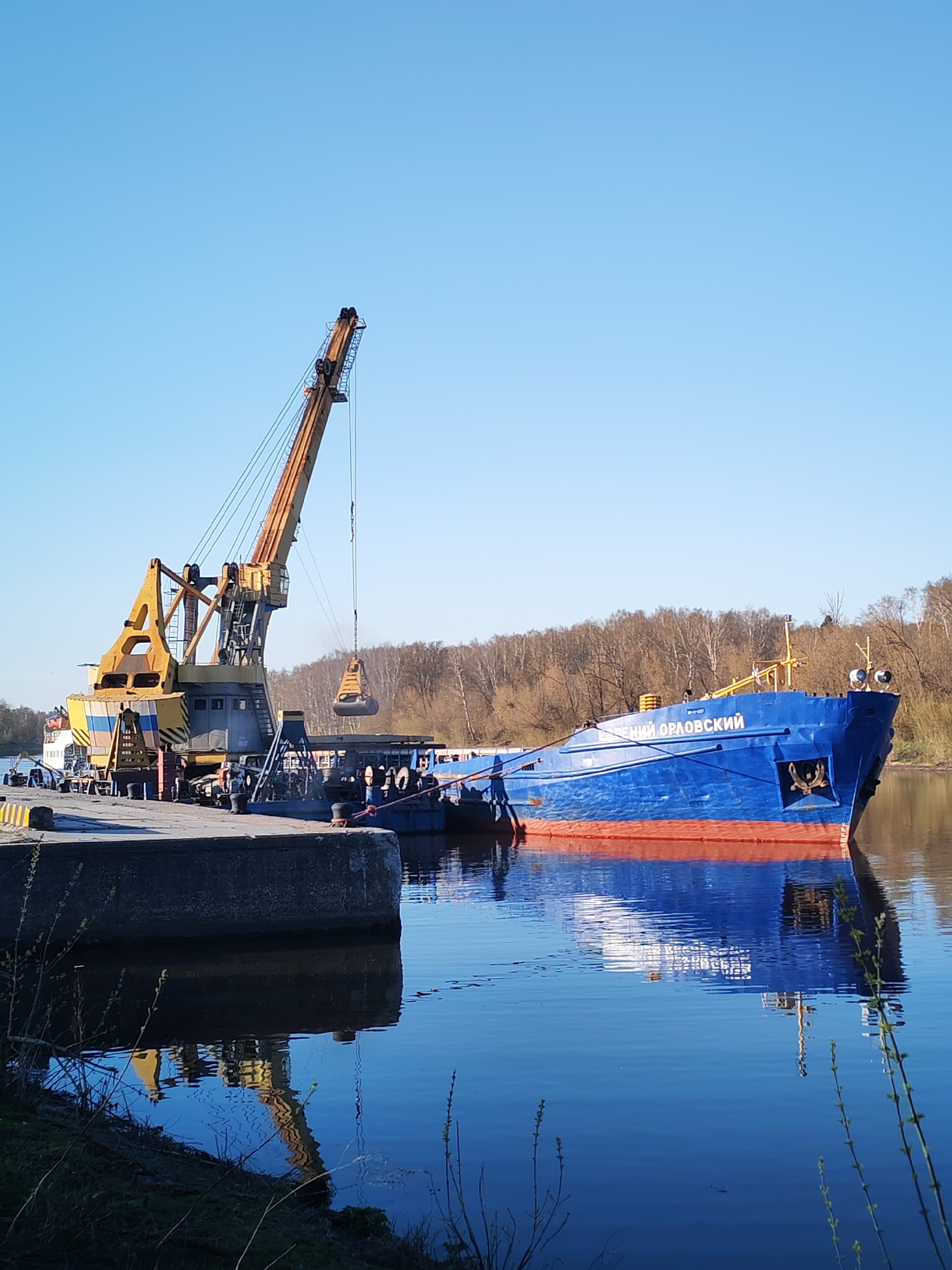 The image depicts a blue and red cargo ship docked at a concrete pier in Yakhroma, Russia. The ship, labeled "Евгений Орловский" (Evgeniy Orlovskiy) in Cyrillic, is being loaded by a yellow and black crane. Several people are standing on the pier near the crane, observing the loading process. The water is calm, reflecting the ship and crane. Bare trees line the opposite bank of the river under a clear blue sky.