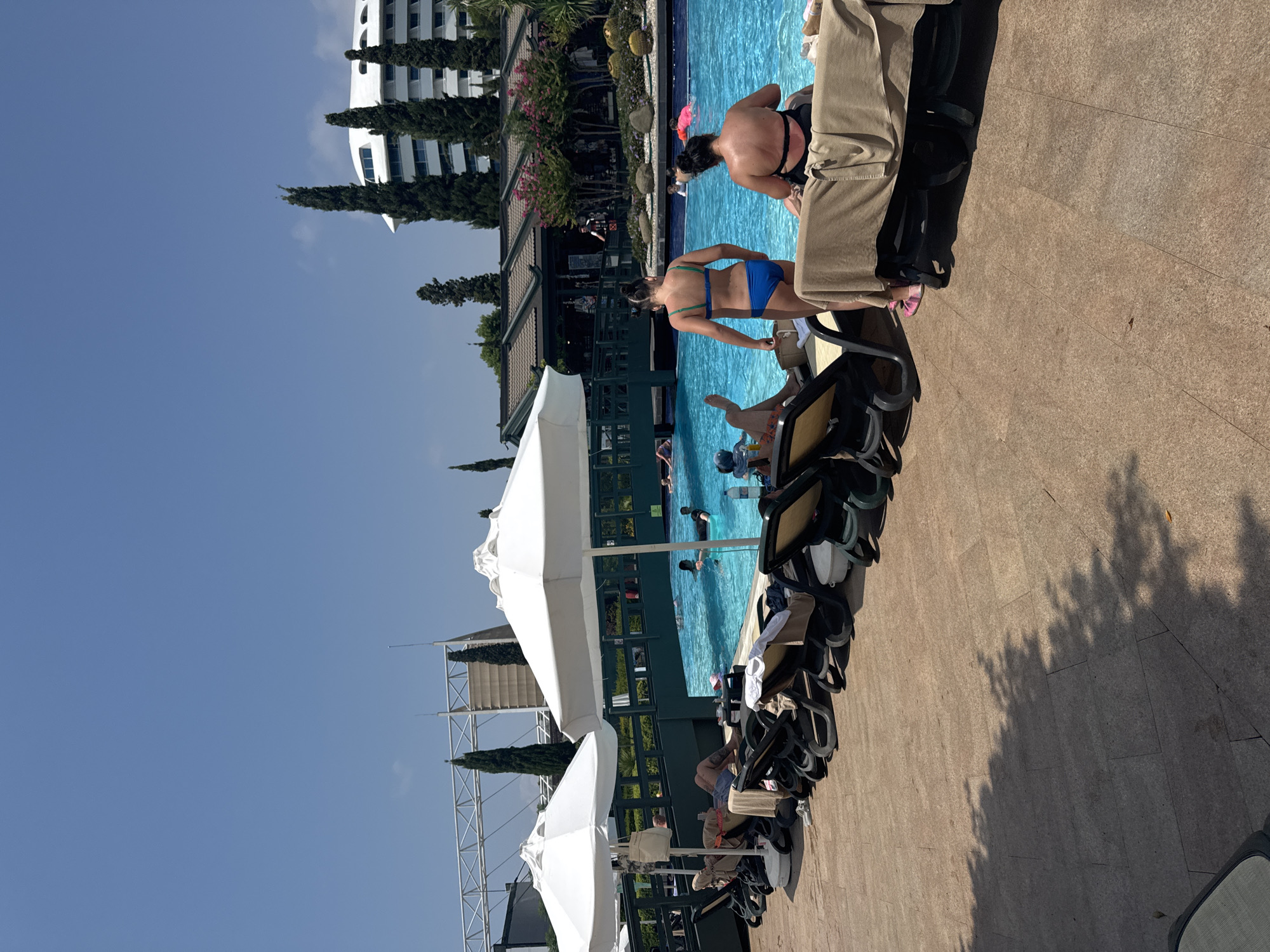 This is a daytime, medium-angle, outdoor shot of a resort pool area in Aksu, Turkey. In the foreground, several empty lounge chairs are set up on a tiled patio, some covered with towels. To the right, two women are seated on lounge chairs. One is wearing a black bikini, the other a blue bikini. Both are facing away from the camera. Beyond the lounge chairs is a large, bright blue swimming pool, where several people are visible swimming and relaxing. A dark green bridge spans across the pool in the mid-ground. Behind the pool, there are buildings with dark roofs and trees, including tall, slender cypress trees. Further in the background, a modern white hotel building with many windows is visible. The sky is clear and blue.