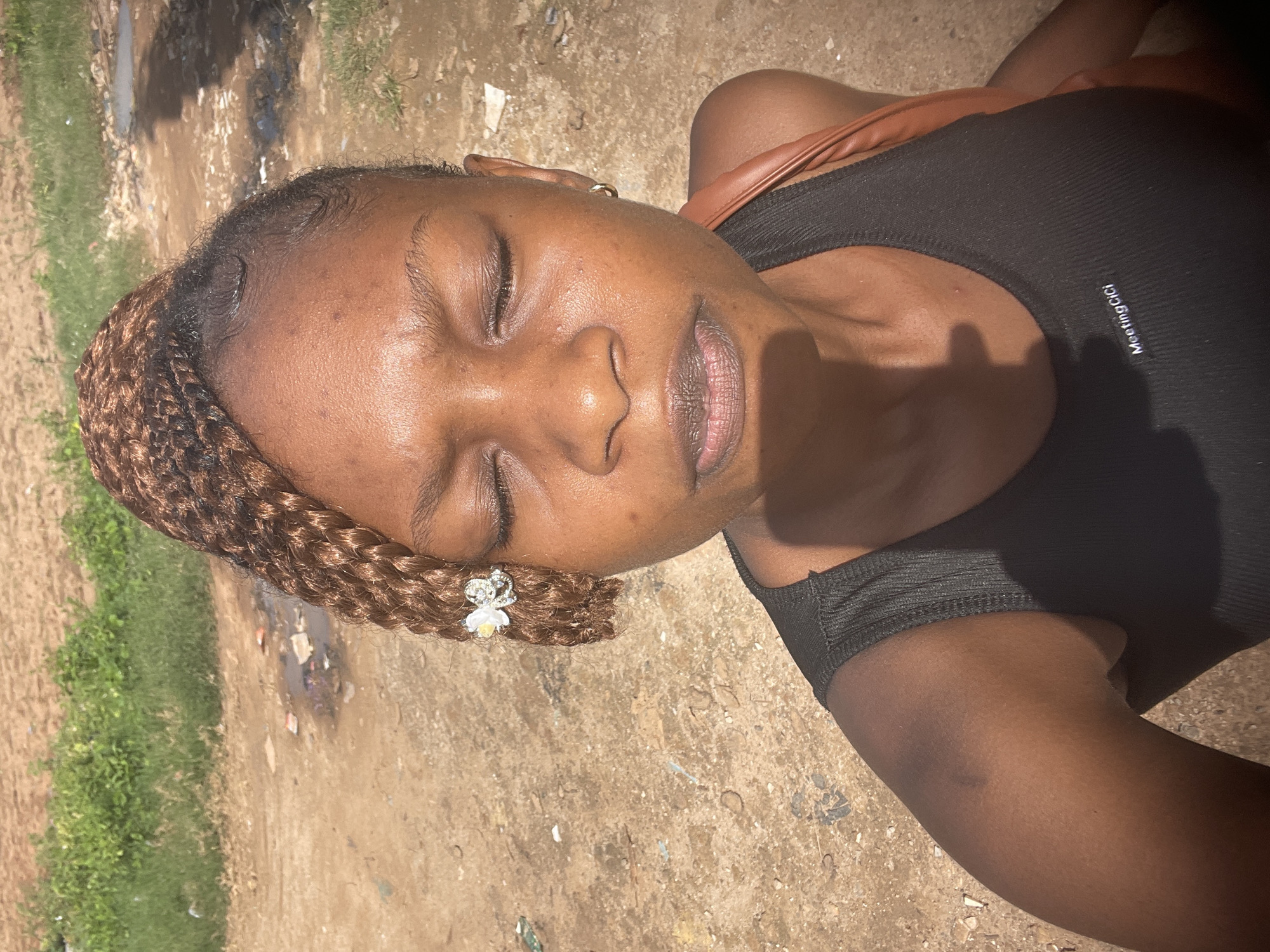 The media file is an image of a young Black woman lying on her back outdoors. She has her eyes closed and appears relaxed. Her hair is braided and adorned with a small, sparkling hair clip shaped like a crown with a flower. She is wearing a black tank top with a thin strap visible over her shoulder, and a brown strap from a bag is also visible. The background is an outdoor scene with a dirt ground, some scattered debris, and patches of green grass. The lighting suggests it is daytime, likely sunny, with shadows visible on the ground and her clothing. There is faint text on her tank top that reads "MeetingGigi". The overall impression is of a serene moment captured outdoors.