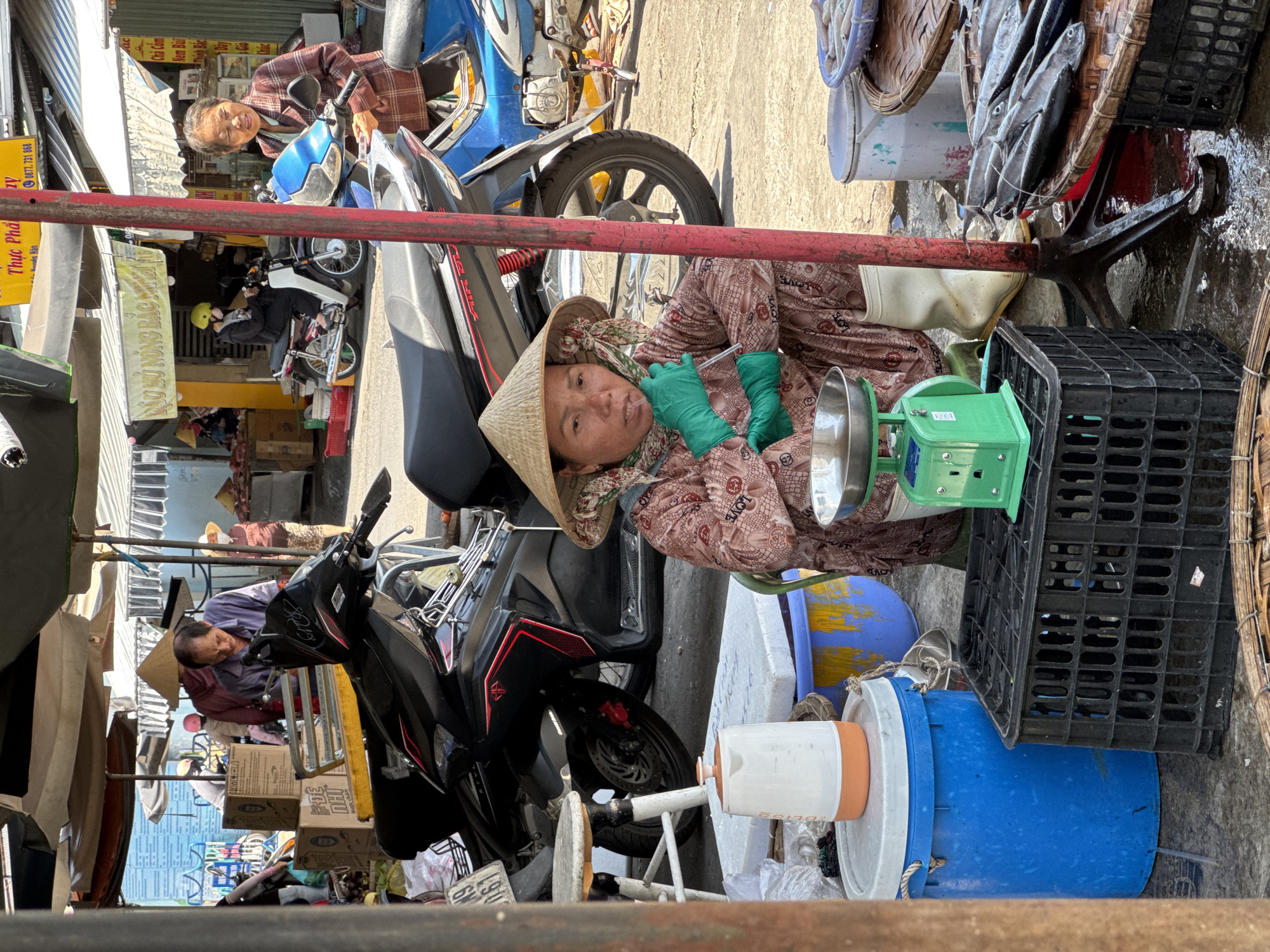 The image captures a street market scene in Nha Trang, Vietnam, likely in the early morning or late afternoon given the lighting and the presence of vendors. A woman wearing a conical hat and green gloves is seated at a stall, operating a green mechanical scale. She is dressed in a patterned shirt and pants. In front of her are various items, including a metal bowl and what appear to be fish or seafood on a woven mat. Behind her, several motorcycles are parked, suggesting a busy thoroughfare or a market where transport is readily available. Other individuals, partially visible, are also present in the background, engaged in market activities. The environment appears to be an open-air market, with awnings providing shade and the ground showing signs of being damp, possibly from washing or recent rain. The overall impression is one of everyday commerce in a vibrant Vietnamese city.