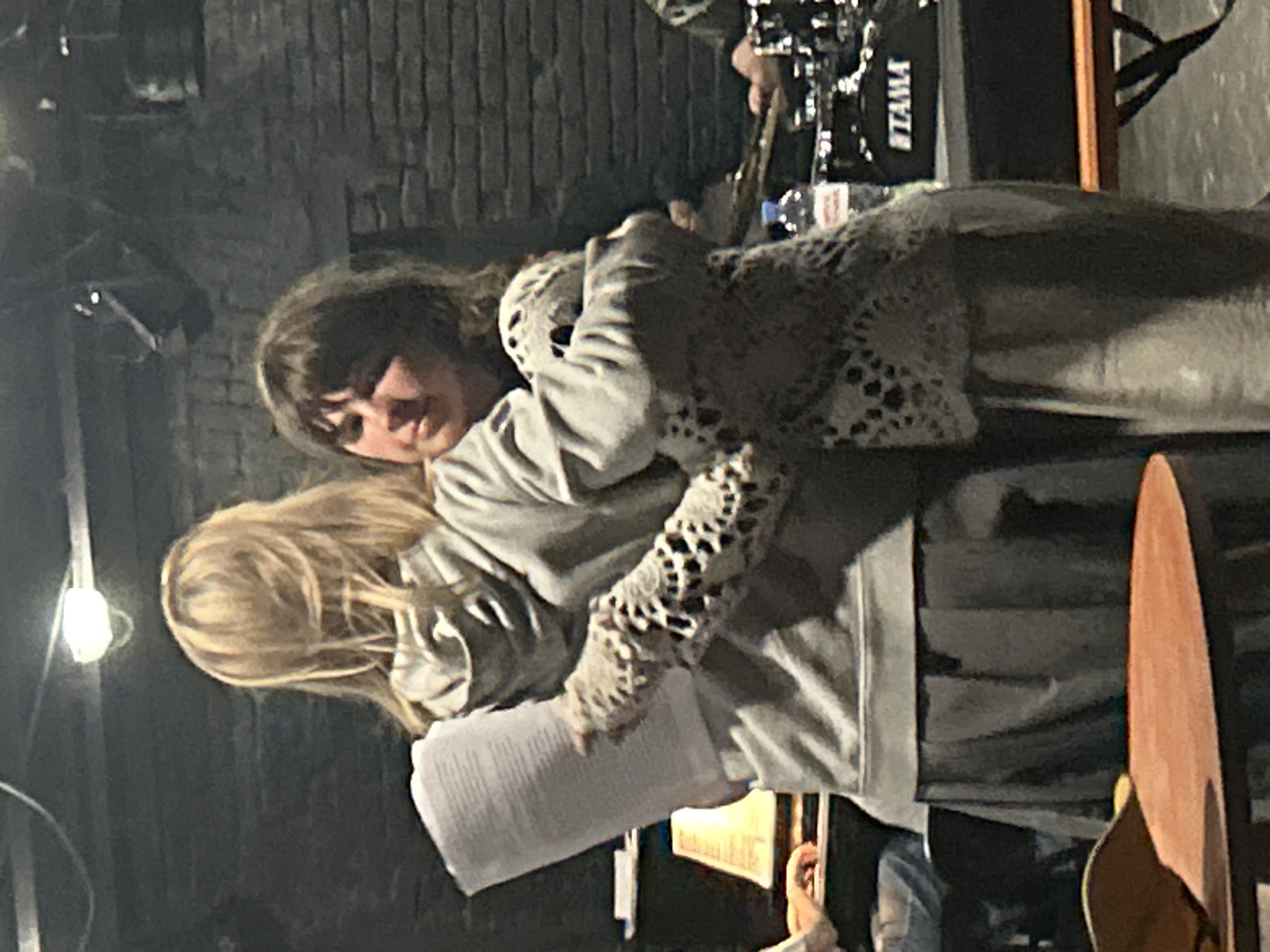 Two women embrace in an indoor setting, near what appears to be a stage area. One woman has brown hair and is looking toward the viewer, while the other has blonde hair and is holding a stack of papers. The women are dressed in casual clothing. Behind them is a partially visible drum set with a "TAMA" logo and what looks like a guitar case. The background suggests a brick wall and possibly a performance venue. The city of Unknown, Unknown cannot be determined from the image.