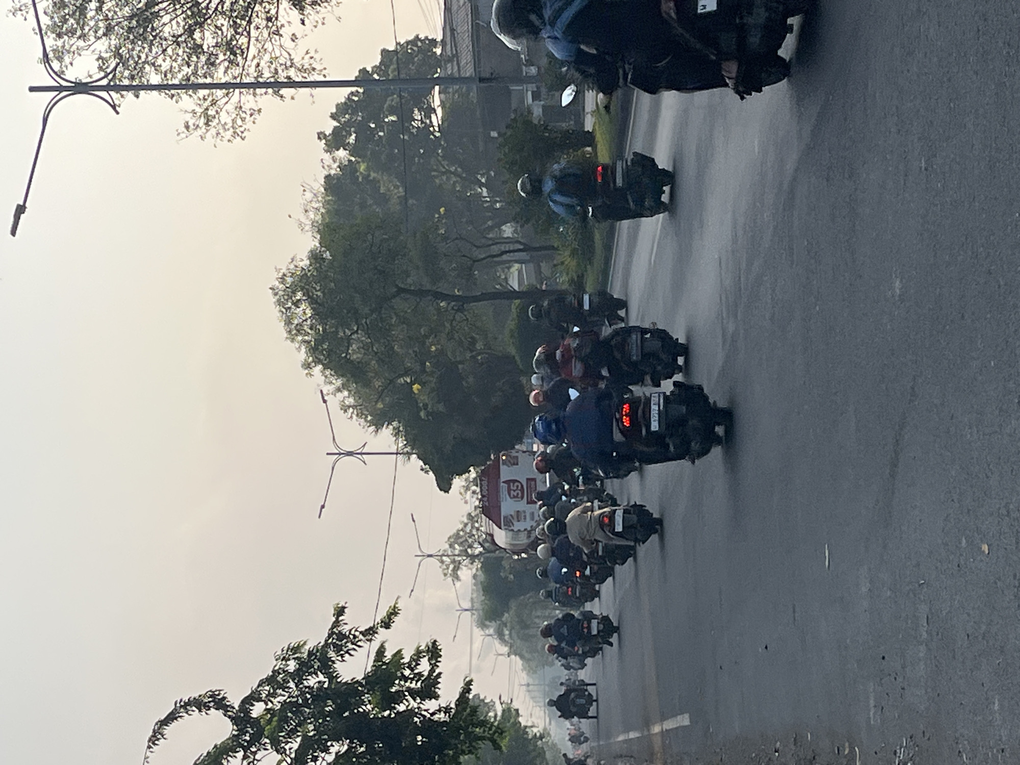 The image shows a street scene in Gelam, Indonesia, characterized by a heavy traffic flow of motorcycles. The perspective is from the side of the road, looking ahead at a convoy of riders on scooters and motorcycles, most of whom are wearing helmets. In the distance, a large, white tanker truck with red lettering, including the number "35", is visible. The road is paved and appears to be a main thoroughfare with streetlights lining the sides. Trees and foliage flank the road, and the sky is hazy, suggesting a warm or polluted environment. The overall impression is one of a bustling urban or suburban commute, typical of Indonesian cities.