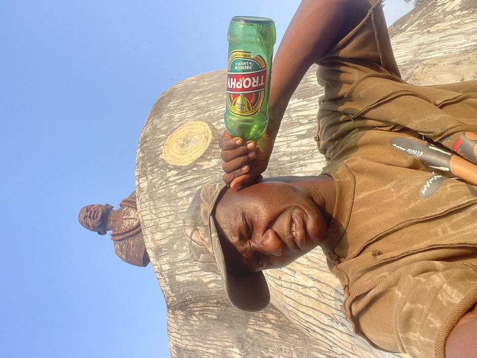 A person in a brown outfit holds a green "Trophy" beer bottle against their forehead in front of a large, grey, elephant-like sculpture with a wood grain pattern. The sky is clear blue. The person wears a camouflage cap and smiles with closed eyes. A small sculpture of a person with a beaded necklace is attached to the side of the larger sculpture. There are also painted objects behind the subject.