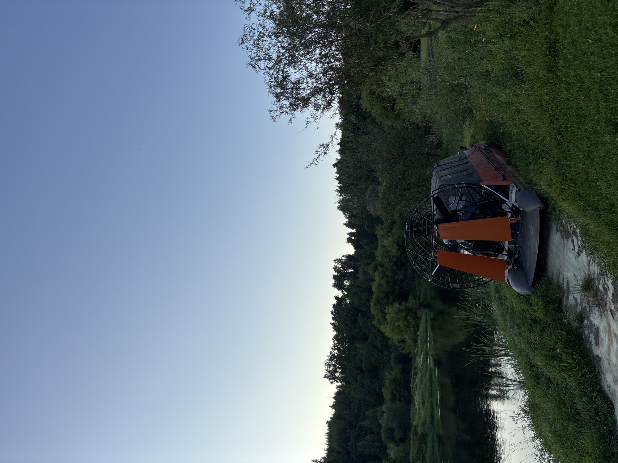 This is a full shot of an airboat docked on the grassy bank of a river. The airboat is red with orange accents and a black cover over the seating area. It is parked on a dirt and grass embankment. The river is calm with dark water reflecting the sky. A dense line of green trees borders the far bank of the river, with a clear, light blue sky above. The image appears to be taken during the day. The location is the riverbank in Zvenigorod, Russia, specifically near the Savvino-Storozhevsky Monastery, a historically significant site.