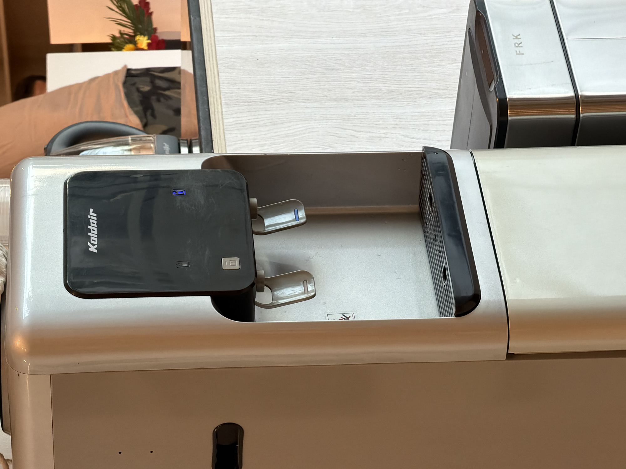 A silver and black water cooler stands to the left of the frame, with a metal trash can labeled "FRK" to its right. The water cooler has two spigots and a black panel with the brand name "Kaldair" and a blue light indicator. Behind the water cooler, a person wearing a tan shirt and green shorts is visible, as well as a kettle and a vase with flowers. The scene appears to be indoors, likely in a public space like an office or hotel lobby. The specific location in Sharm El Sheikh, Egypt, cannot be determined from the provided image.