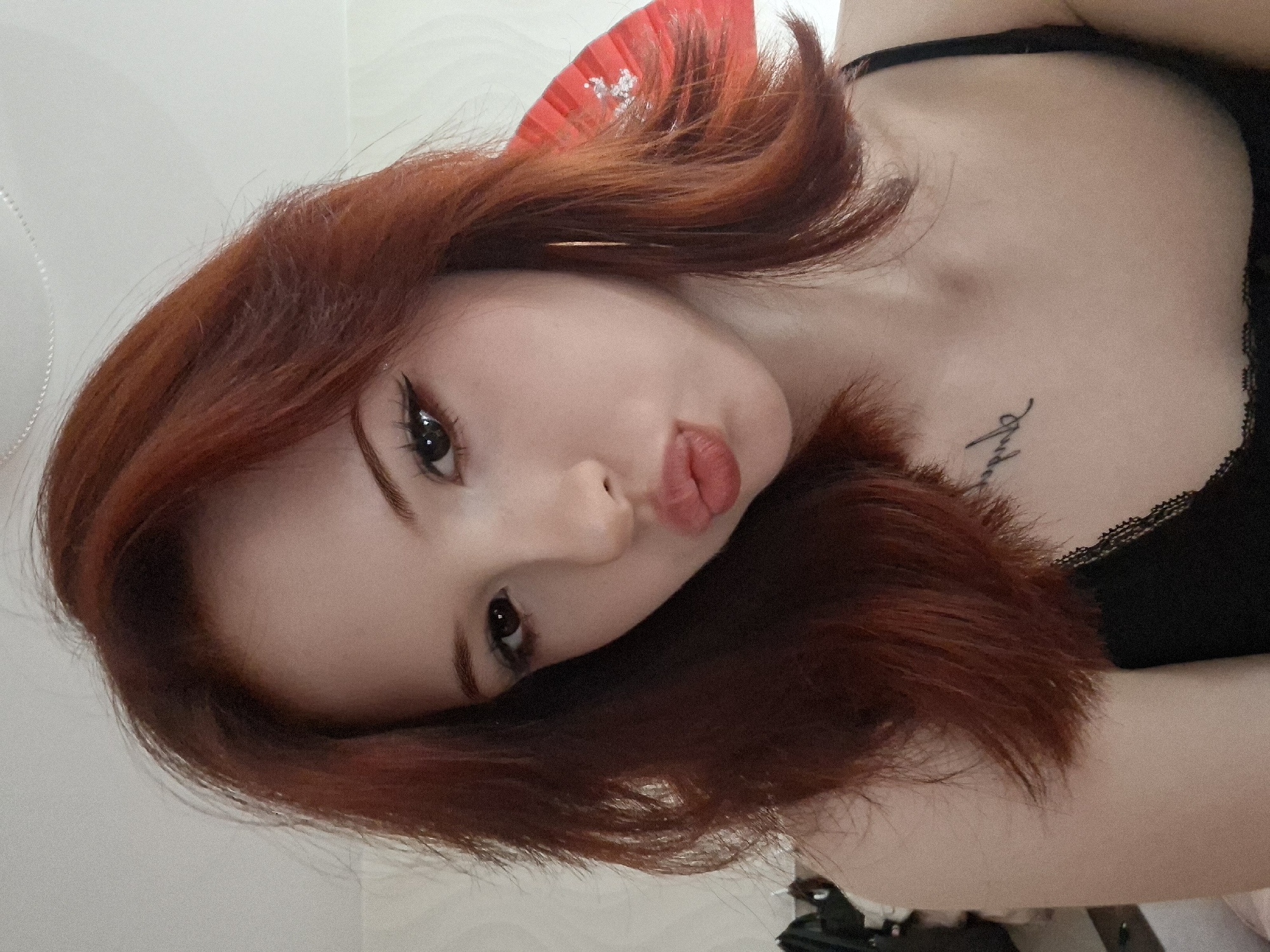 A young woman with red hair is lying down, facing the camera. She has fair skin, dark eyes with winged eyeliner, and is making a kissing face. She is wearing a black top with lace trim. A decorative red fan is visible in the background, partially obscured by her hair. A tattoo is visible on her upper chest. The background appears to be a plain white or light-colored wall. It is unknown if this image was taken in Chaikovsky, Russia.