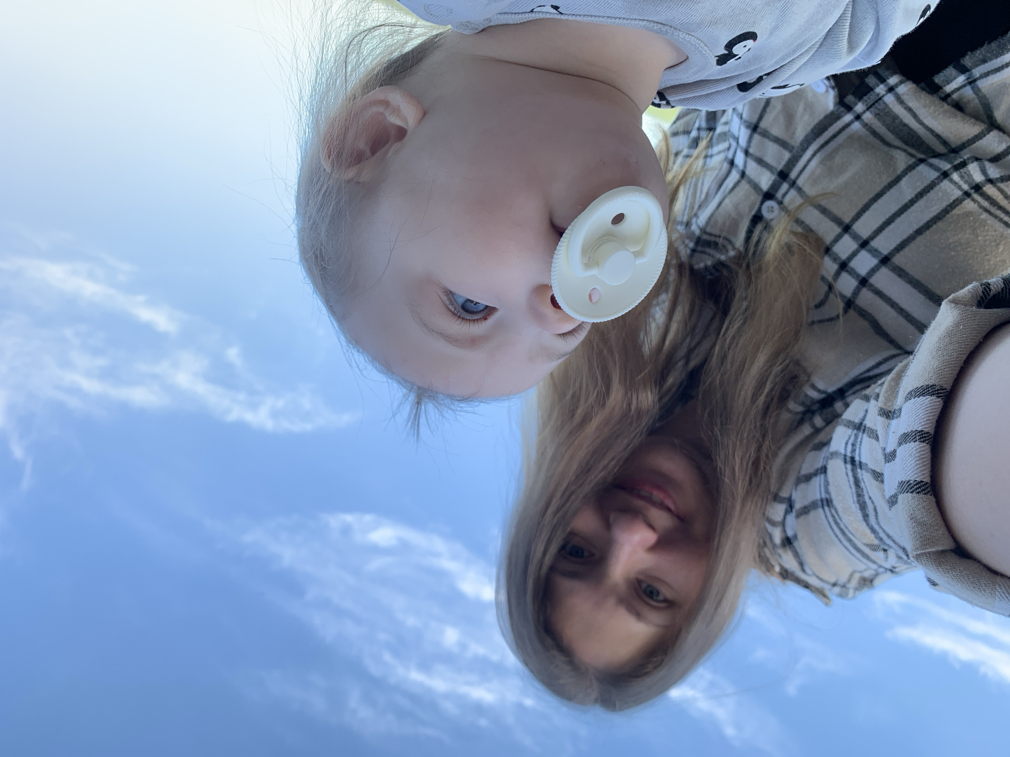 A low-angle photograph shows a woman and a baby with a pacifier outdoors, against a backdrop of a blue sky with light clouds. The woman has long brown hair and is wearing a plaid shirt. The baby is wearing a light-colored shirt with cartoon images. The image's upside-down orientation makes it difficult to definitively establish a location in Chaikovsky, Russia, as the environment is largely obscured.