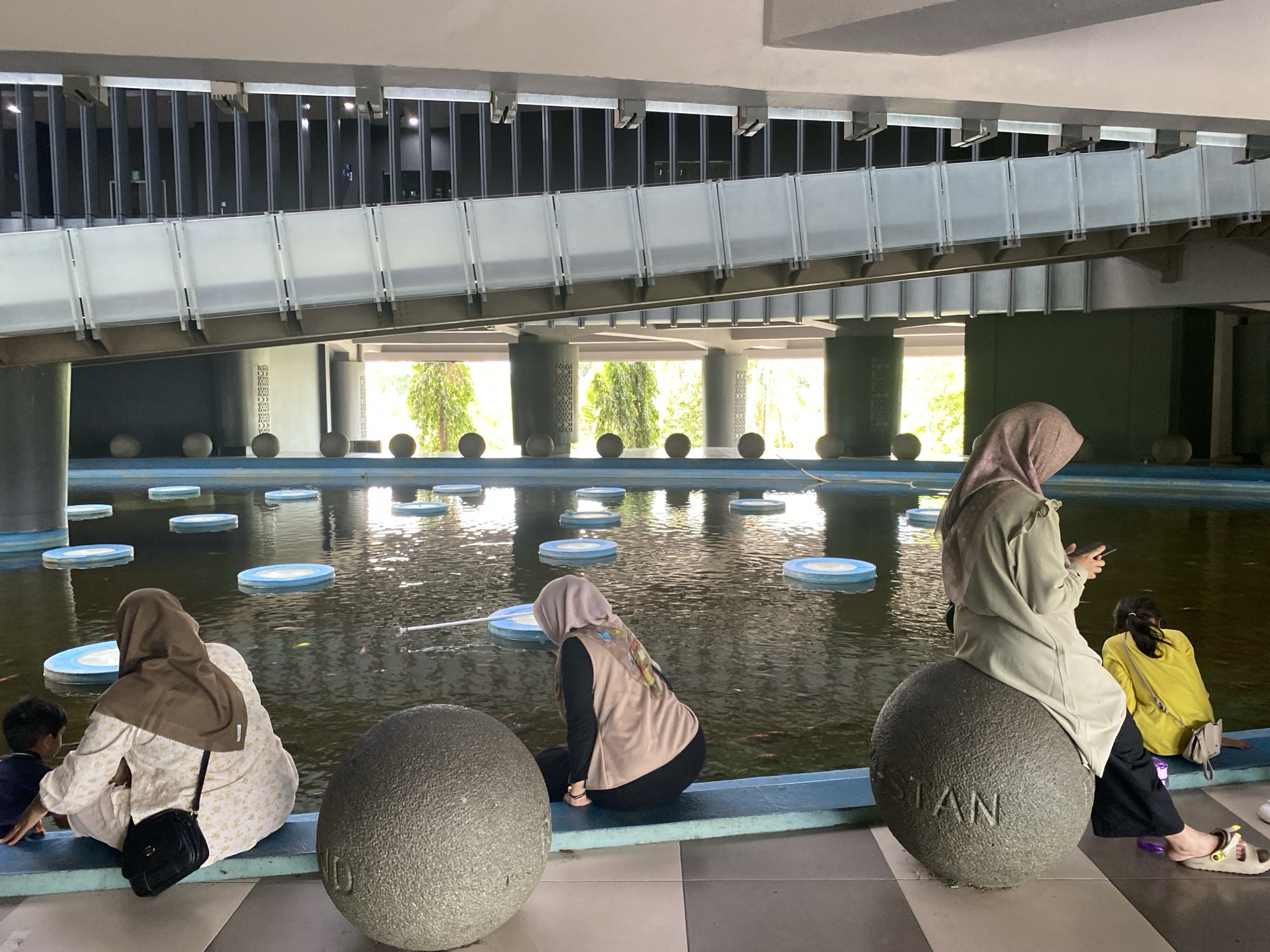 In Banda Aceh, Indonesia, a photograph captures a serene outdoor scene beneath a modern, elevated walkway. The walkway features a sleek, metallic structure with frosted glass panels and visible supports. Below, a manmade pond or water feature dominates the foreground, dotted with circular, blue-rimmed platforms. The water is dark and reflects the ambient light, with several small fish visible beneath the surface.
Three women and one young child are present, all appearing to be of Southeast Asian descent. Two women, dressed in modest Islamic attire, sit on the edge of the pond. One, wearing a beige hijab and a floral pattern dress, is crouched down, seemingly interacting with the child, who is wearing a dark blue shirt and is partially visible on the left. A black crossbody bag rests on the woman's shoulder.
Another woman, in a light pink hijab and a tan vest over a black long-sleeved shirt and pants, sits on a large, spherical, stone-like object facing the pond. Her hands are occupied with her smartphone. A young girl, wearing a bright yellow shirt, sits beside her on a similar stone object, facing away from the camera.
The third woman, also wearing a hijab, is seated on a large, spherical stone object with the word "STAN" inscribed on its surface. She is dressed in a light olive-green outer garment and dark pants, and she is holding a smartphone, seemingly texting or browsing.
The environment suggests a public space, possibly a park or a cultural complex, with a paved area in the foreground and greenery visible beyond the pond. The lighting indicates it is daytime, with soft sunlight filtering through the structures above and the trees in the background. The overall atmosphere is tranquil and contemplative.
