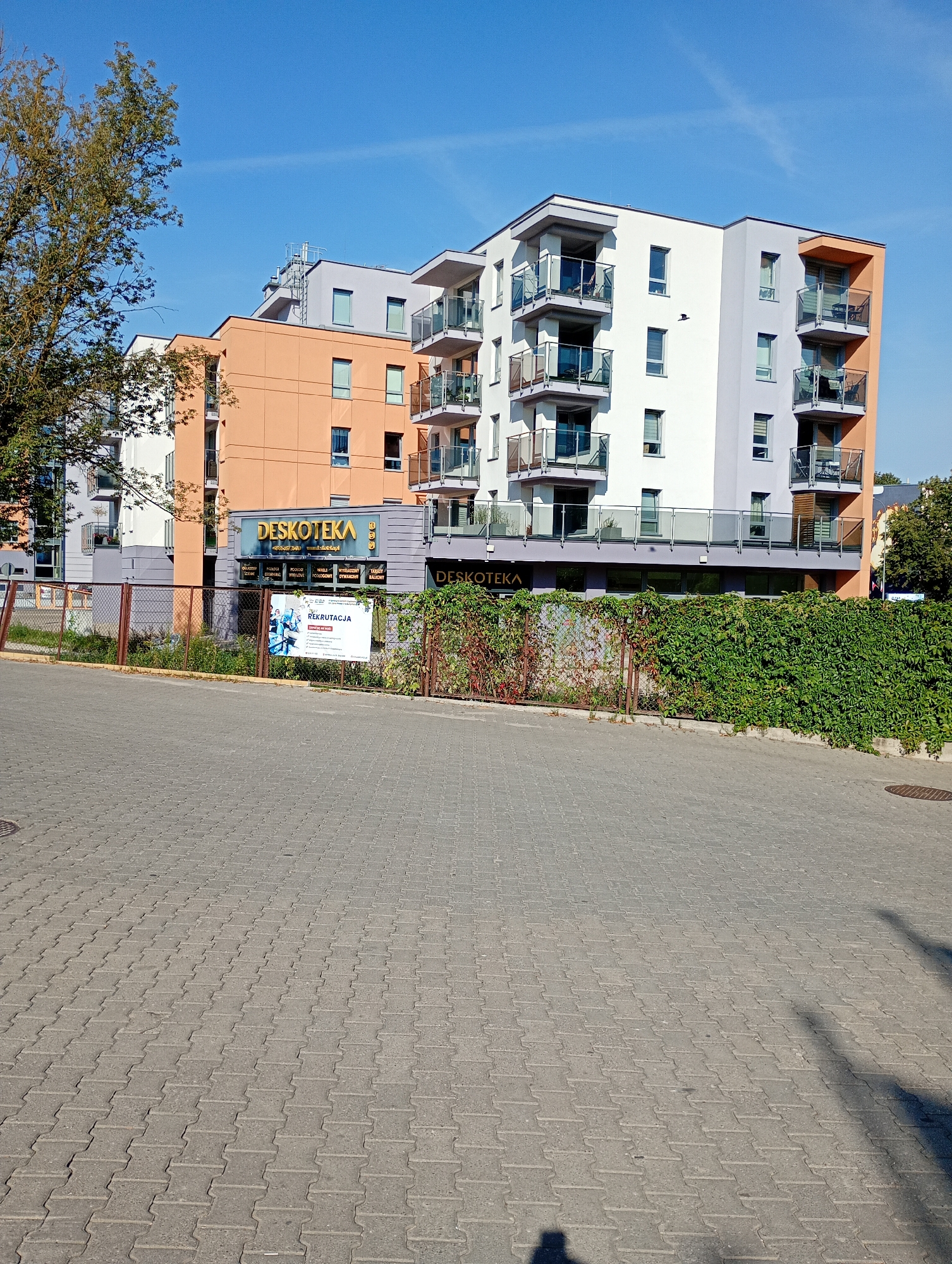 The image depicts a modern apartment building with a mixed facade of orange, white, and gray sections. The building features multiple balconies with glass railings. A sign for "DESKOTEKA" is visible on the ground floor, along with a smaller sign advertising "REKRUTACJA" (recruitment). The building is situated in Białystok, Poland, as indicated by the presence of text on the signs and the overall architectural style common in Polish cities. The foreground shows a paved walkway with interlocking concrete pavers, and a chain-link fence covered in green ivy separates the walkway from the building's grounds. The sky is clear and blue, suggesting a sunny day. There are no people clearly visible in the image, but shadows on the pavement suggest the presence of at least one person off-camera. A tree with green leaves stands to the left of the building.