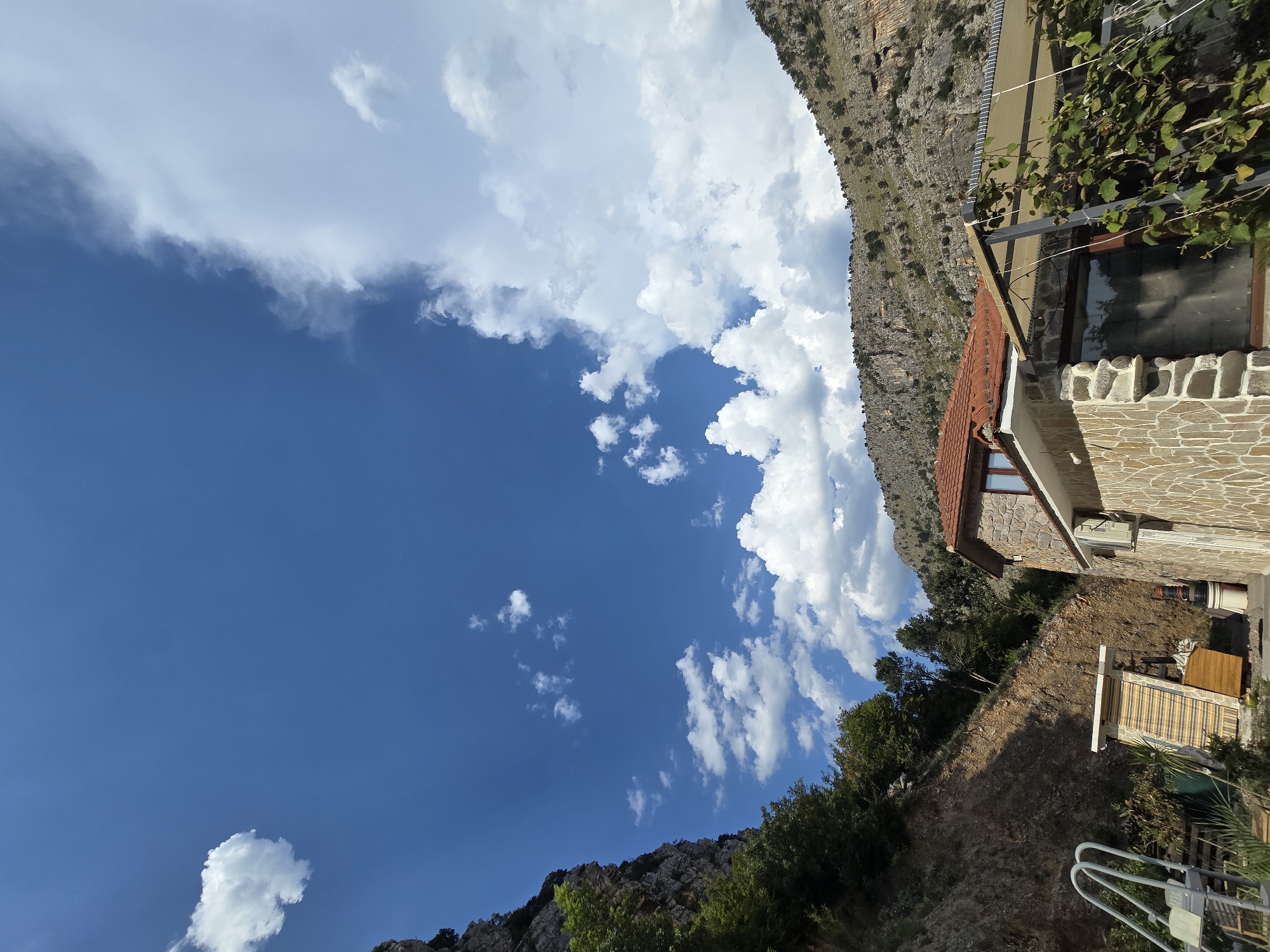 The image depicts a residential building with a stone facade and a red tile roof, situated in a mountainous area. A portion of a steep, rocky slope covered in vegetation is visible to the left. In the foreground, there is an outdoor seating area with a canopy, and some climbing plants. To the left of the building, a small, utility structure with a striped awning is visible. The sky is bright blue with scattered white clouds, and the overall impression is of a rural or semi-rural setting, likely in a mountainous region. Based on the context, the location is identified as Podgorica, Montenegro, a city characterized by its proximity to mountainous terrain and scenic landscapes.