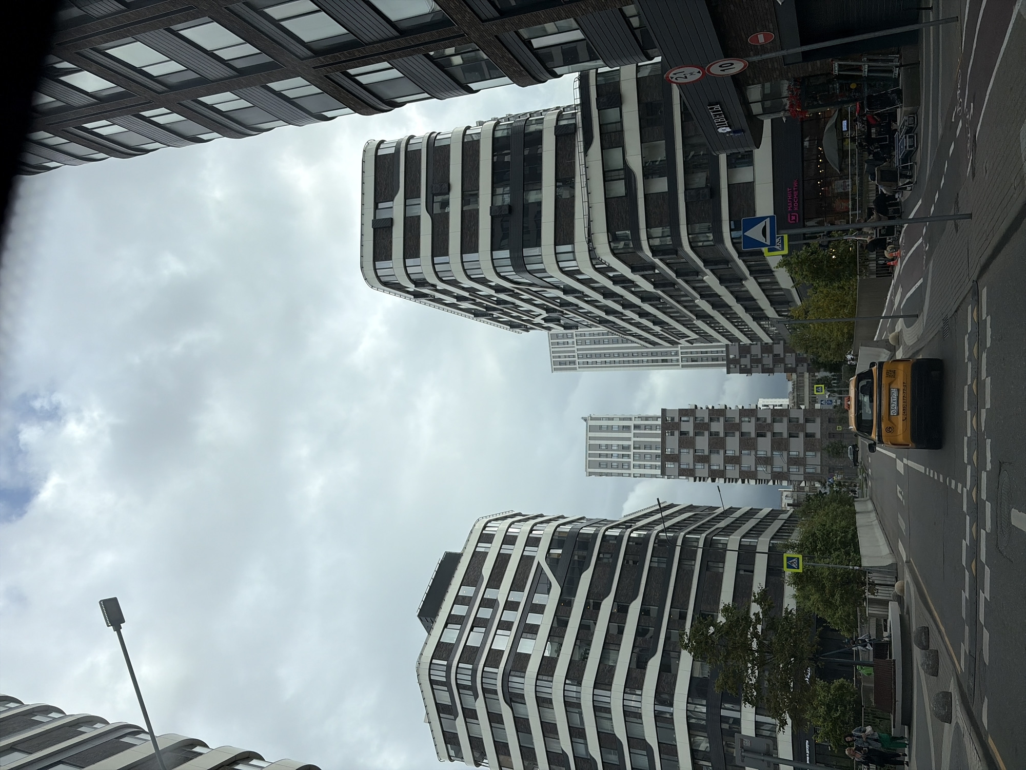 The image captures a street view in Moscow, Russia, characterized by modern, high-rise residential buildings. The architecture features a blend of dark brick facades and white trim around numerous windows, creating a distinctive striped pattern on the buildings. Several of these contemporary structures are visible, dominating the skyline under a cloudy sky.
In the foreground, a yellow taxi is driving down the street. The road has visible lane markings and pedestrian crossings. Along the sides of the road, there are trees, sidewalks, and what appear to be commercial establishments at the ground level of some buildings, with signage indicating shops like "Магнит Косметик" (Magnit Cosmetic) and "ЦВЕТЫ" (Flowers).
Streetlights and traffic signs are also present, including a sign with a red circle and the number "10," likely indicating a speed limit, and another with a red circle and a horizontal bar, possibly a "no entry" sign. There are a few people walking on the sidewalk and near the buildings. The overall impression is of a developed urban environment with a focus on modern architecture.