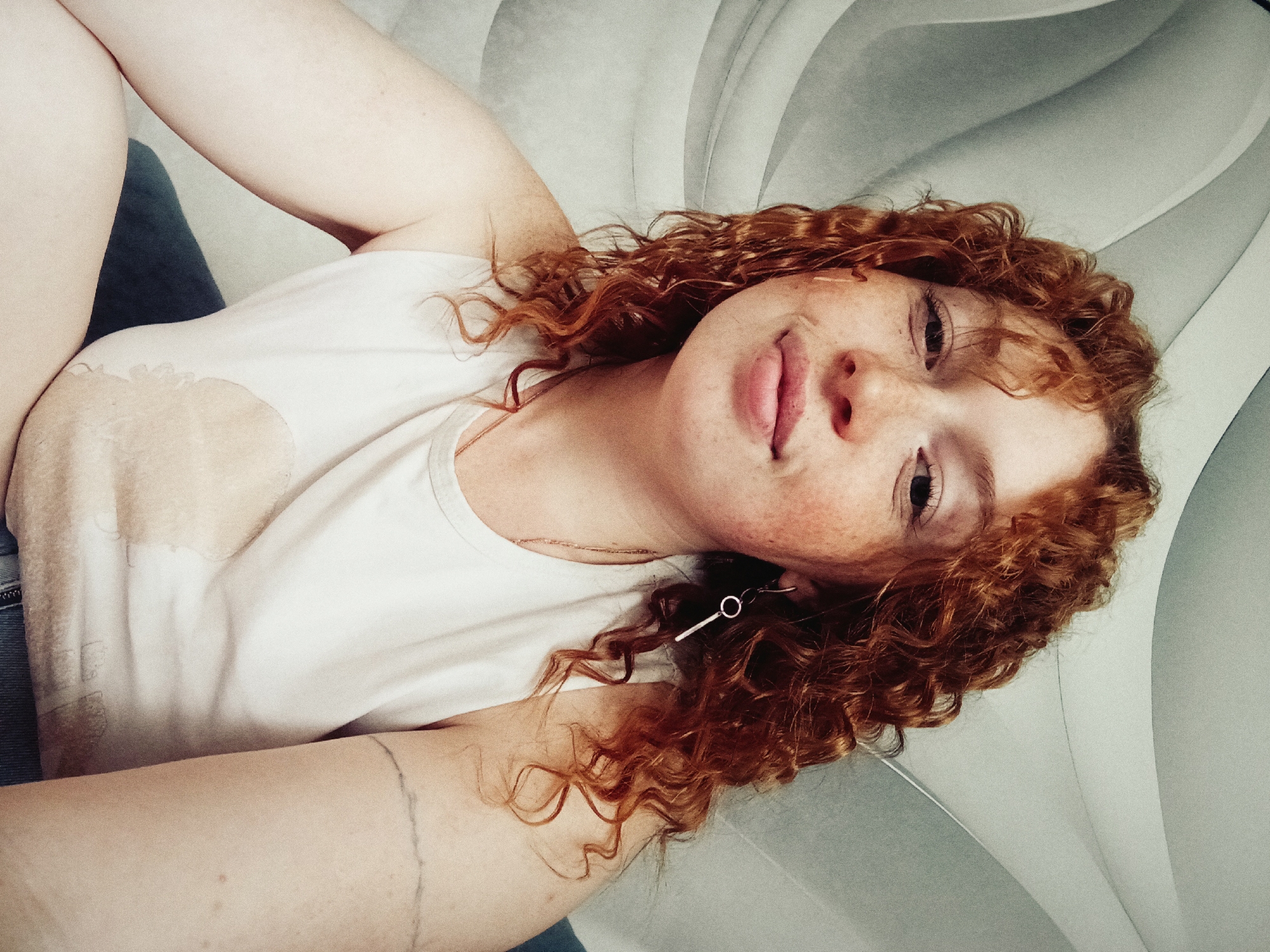 This is a close-up, high-angle portrait of a young adult woman lying down and looking directly at the camera. She has fair skin, a scattering of prominent freckles across her nose and cheeks, and voluminous, curly, reddish-brown hair. Her expression is soft and calm, with a slight, gentle smile and her lips slightly parted.
She is wearing a simple white tank top or t-shirt, and a thin gold-colored necklace is visible around her neck. A distinctive silver-toned earring, composed of a small circular hoop connected to a long, slender bar, hangs from her right earlobe (viewer's left). A dark blue fabric, possibly denim, can be seen near her left side, suggesting she is wearing jeans or similar attire.
The background is an indistinct indoor setting, featuring a light gray or off-white surface with gentle, curved contours, possibly a patterned headboard, sofa, or upholstered furniture. The lighting is soft and even, suggesting ambient light rather than direct sunlight. No specific time of day or weather conditions are discernible from the image. The overall mood is relaxed and intimate. No text is visible. The provided location context for this media file is Yurgamysh, Russia.