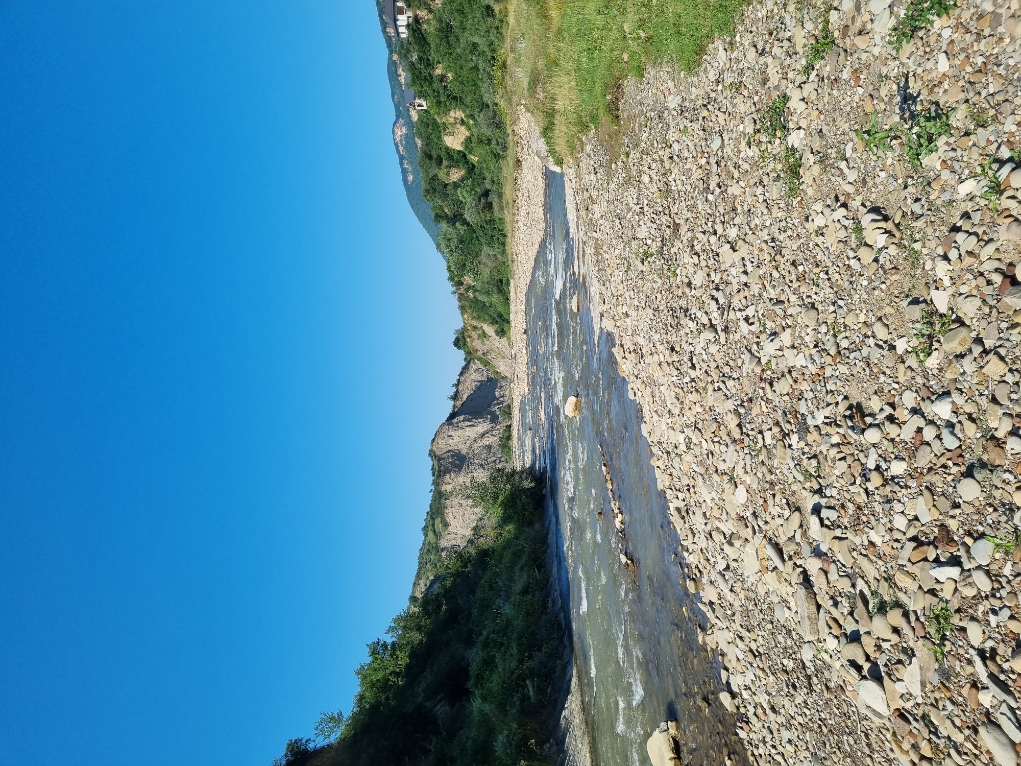 The image depicts a wide river flowing through a rocky valley under a clear blue sky. The foreground is dominated by a dense collection of smooth, rounded stones of various sizes, forming a natural bank. The river itself is a vibrant blue-green, with visible currents and white foam indicating its flow. On the left bank, steep, eroded hills rise, covered in lush green vegetation and trees. In the distance, the valley continues, with more hills and the river winding its way through. On the right bank, the terrain transitions to a grassy slope dotted with more rocks. Two buildings are visible in the upper right portion of the image, nestled on a hillside. This scene is likely located near Khuchni, Russia, a region known for its natural beauty and river valleys.