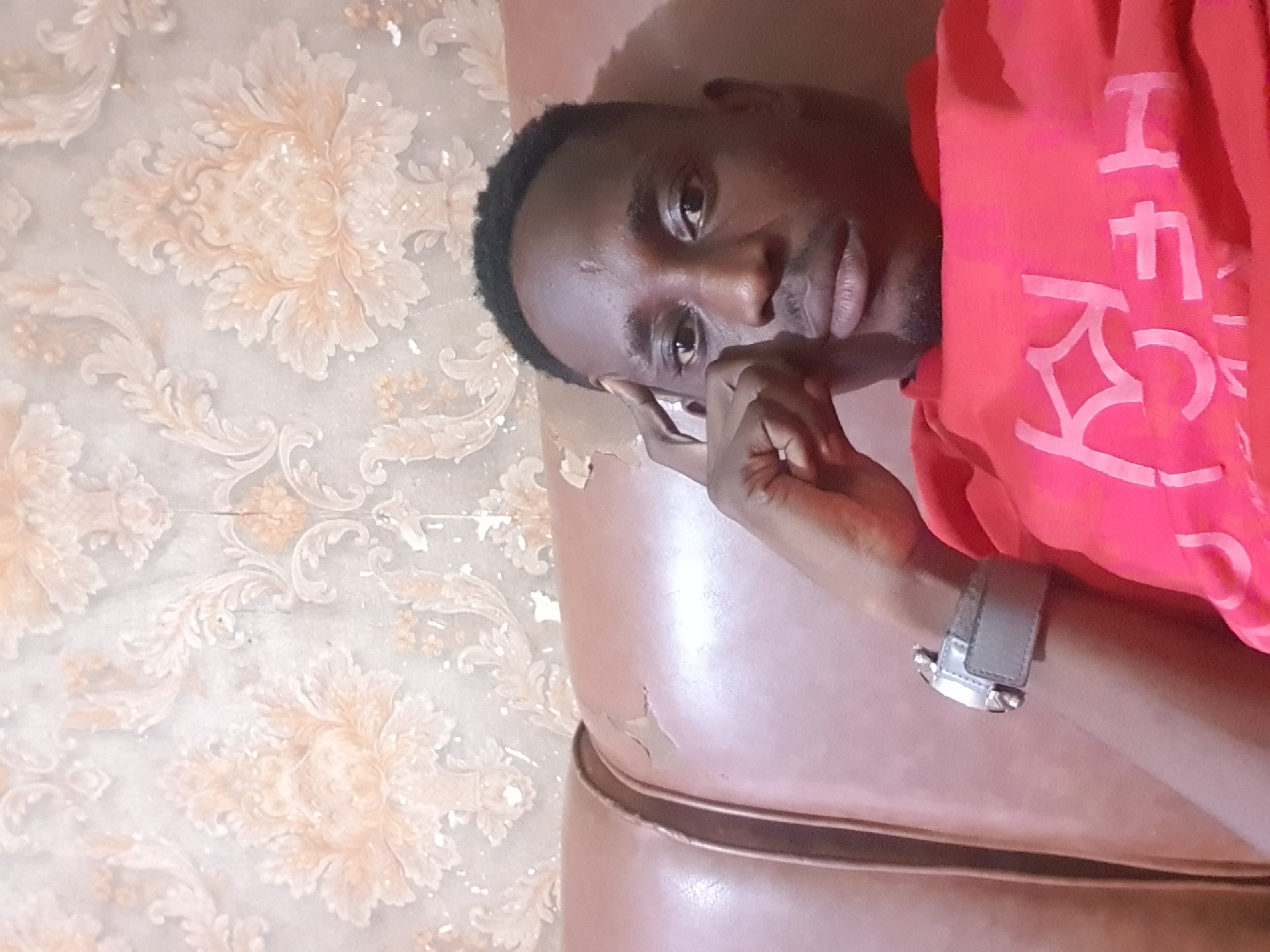 A close-up, head-and-shoulders selfie taken from a low angle shows a Black man with dark skin and short black hair resting his head on his left hand, which is propped up by his index finger and thumb. His right hand is not visible. He is wearing a bright red t-shirt with a large, stylized white graphic on the chest. A silver-colored wristwatch with a grey strap is visible on his left wrist.
The man is seated on a brown leather couch, which appears worn and slightly cracked in places. Behind him, the wall is covered in a light-colored, ornate wallpaper featuring a repeating floral or damask pattern in pale peach and cream tones. The lighting is somewhat dim, casting soft shadows. The overall impression is that of a casual, indoor setting. Based on the provided information, this scene is located in Oluawo, Nigeria.
