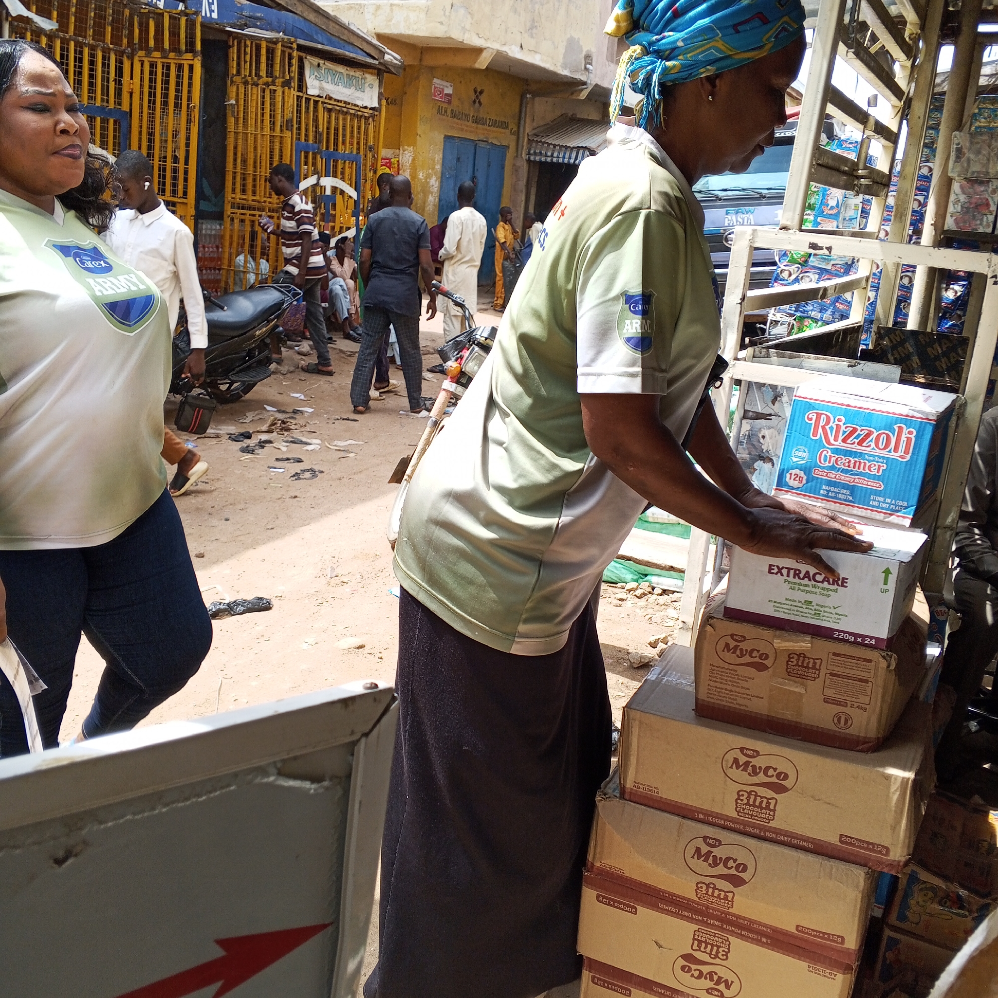 The image depicts a street scene in Arawa, Nigeria, focusing on a woman in the foreground wearing a light green t-shirt with a shield logo and a blue headscarf. She is standing near a motorcycle and a stack of cardboard boxes, one of which is labeled "Rizzoli Creamer." Behind her, other individuals are visible, including a man in a striped shirt and several others in the background amidst stalls and buildings. The ground is dusty and littered with debris. The overall environment suggests a bustling market or street-side commerce.