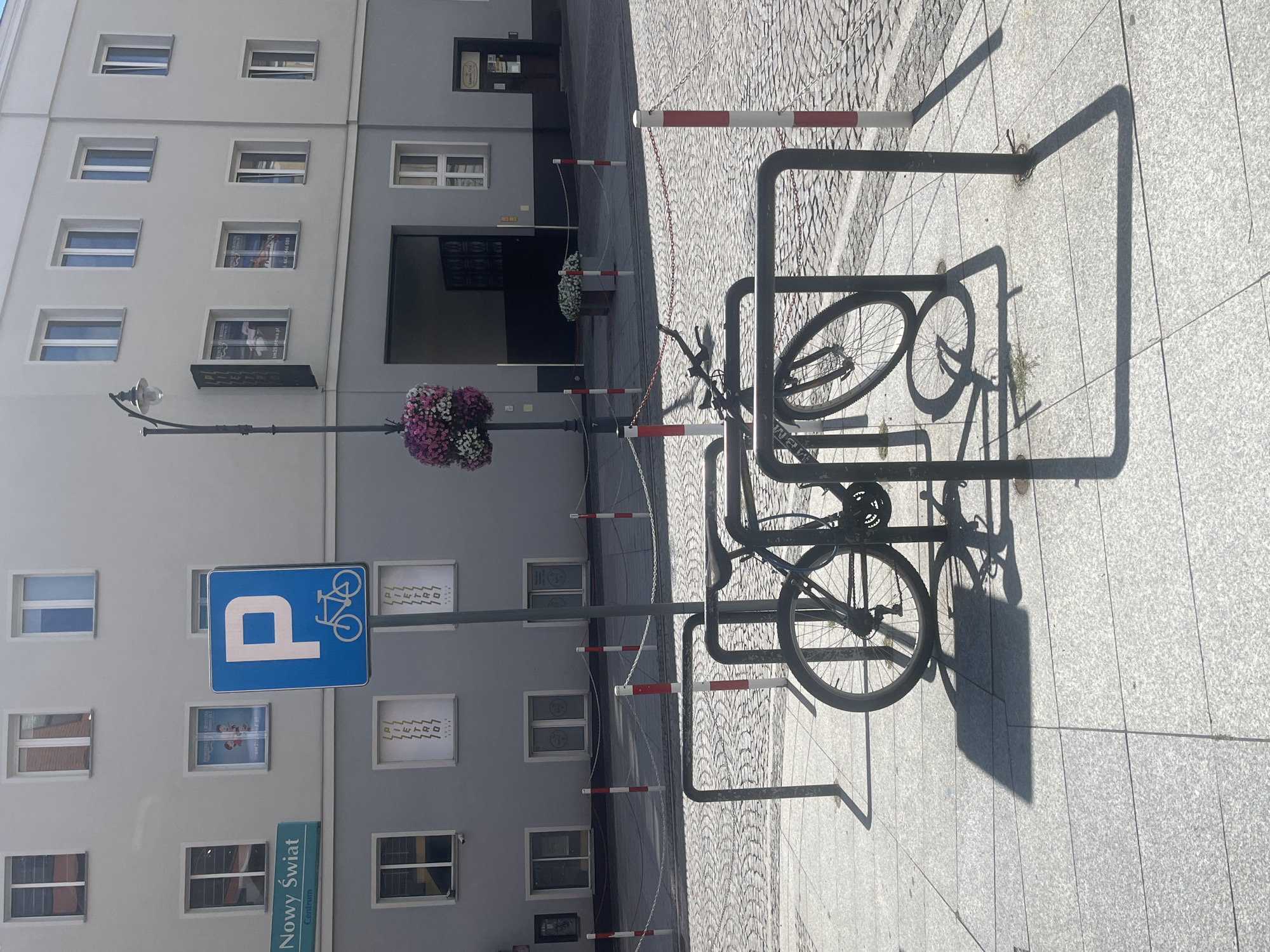 The image shows a street scene in Białystok, Poland, specifically on Rynek Kościuszki, near the Branicki Palace. In the foreground, there is a bicycle rack with two bicycles parked. The bicycles are dark-colored and appear to be standard adult bicycles. The bicycle rack is made of black metal and is designed to hold multiple bicycles.
Behind the bicycle rack, there is a cobblestone street. The street is bordered by a sidewalk with light-colored paving stones. A lamppost with a decorative element of pink and white flowers is visible to the left of the frame. A blue parking sign with a bicycle symbol is mounted on a pole near the lamppost.
In the background, a building with light-colored walls and multiple windows is depicted. Some of the windows have advertisements or posters displayed inside. The building has a gray lower section. The overall impression is a sunny day in an urban environment.