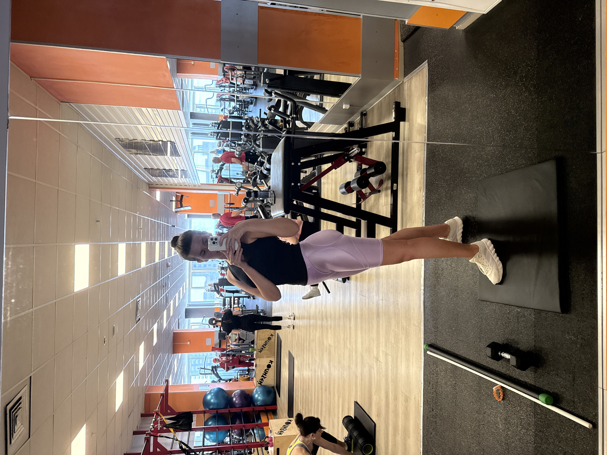 This image captures a bright, active gym interior, likely during the daytime, in Krasnoyarsk, Russia. The primary subject is a woman taking a mirror selfie. She is wearing a black tank top, light purple biker shorts, and white sneakers, with her dark hair pulled back into a bun. She holds a white smartphone to capture her reflection in a large wall-mounted mirror.
The gym is well-lit by both rectangular ceiling lights and natural light streaming through expansive windows in the background, which reveal an urban landscape. The flooring consists of both light wooden panels and black speckled rubber mats. Orange accent panels on some walls add a touch of color to the otherwise neutral ceiling and wall palette.
In the reflected background, several other individuals are visible. Some people in red shirts appear to be using weight machines. Another person, dressed in black, is reflected further away. In the bottom right foreground (when viewed upright), a woman in a yellow and green top is engaged in stretching or foam rolling on a black mat. Adjacent to her on the rubber floor are a black foam roller, a black dumbbell, a white and green exercise bar, and a small orange bracelet. A rack holds several blue exercise balls, and some wooden boxes labeled "KOUNYEN" are also present, adding to the typical gym setting. The scene depicts a lively fitness environment with multiple individuals engaged in various workouts.
