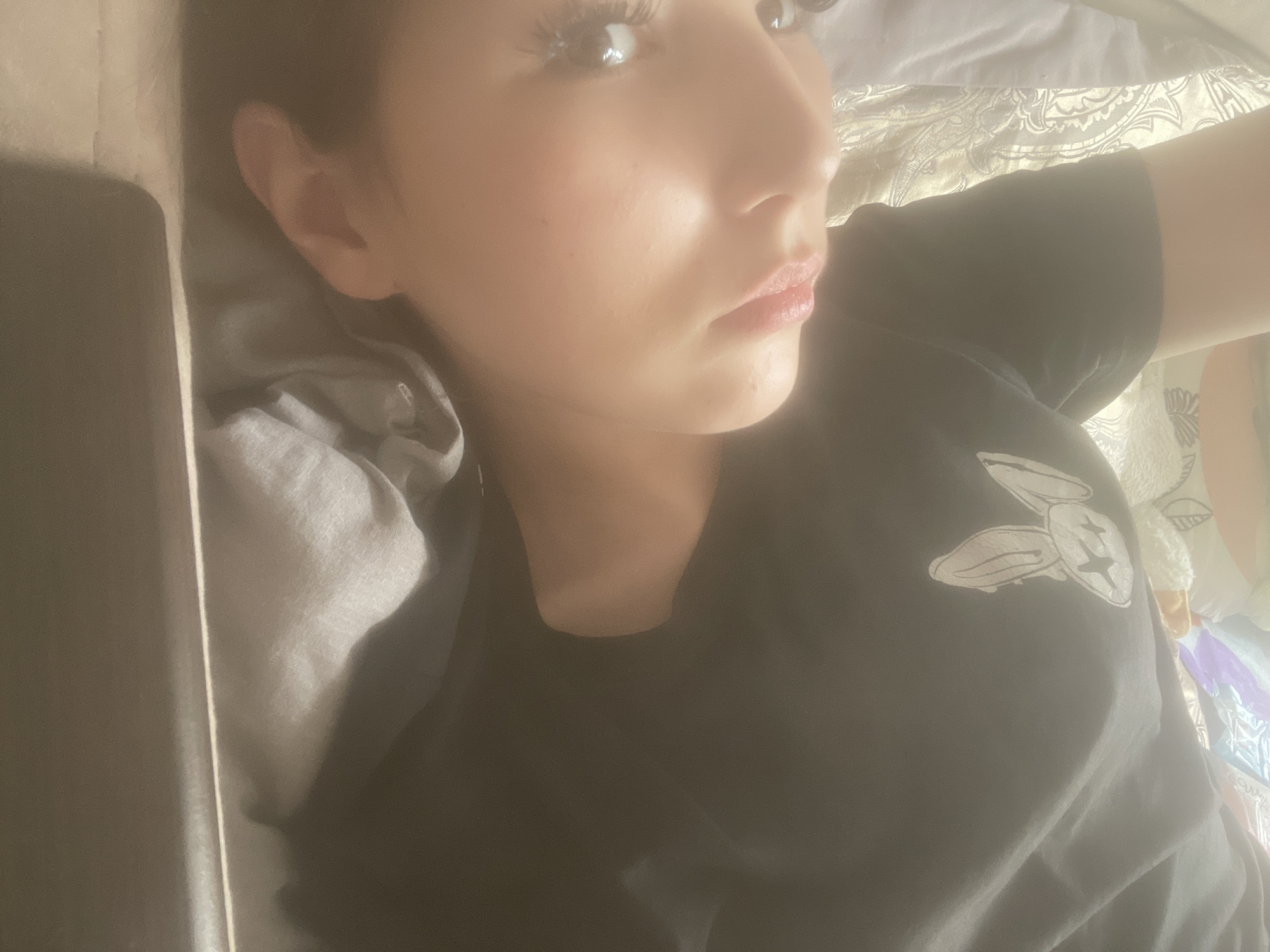 A close-up, hazy image captures a person, likely female, lying down indoors, possibly in a bed. The individual is seen from the chest up, positioned on their side, with their gaze directed upwards and to the right, away from the camera. They have visible eyelashes and glossy lips. They are wearing a dark, possibly black, t-shirt featuring a white graphic on the upper chest. The graphic consists of three stylized shapes resembling abstract bunny heads or skulls, one of which contains a cross, hinting at the "Bad Bunny" logo.
The person rests their head on a grey pillow or bedding. Behind their back and arm, a lighter-colored fabric with a patterned design, possibly damask or paisley, is visible. To the left of their head, a dark, rectangular object, such as a book, tablet, or phone, is partially obscured. The overall lighting is soft and diffused, lending a slightly ethereal or dreamy quality to the scene. The activity suggests resting or quiet observation. The time of day is not explicitly clear, but the gentle illumination implies indirect light. No text is visible in the image itself. The location context provided is Sinyavino, Russia.