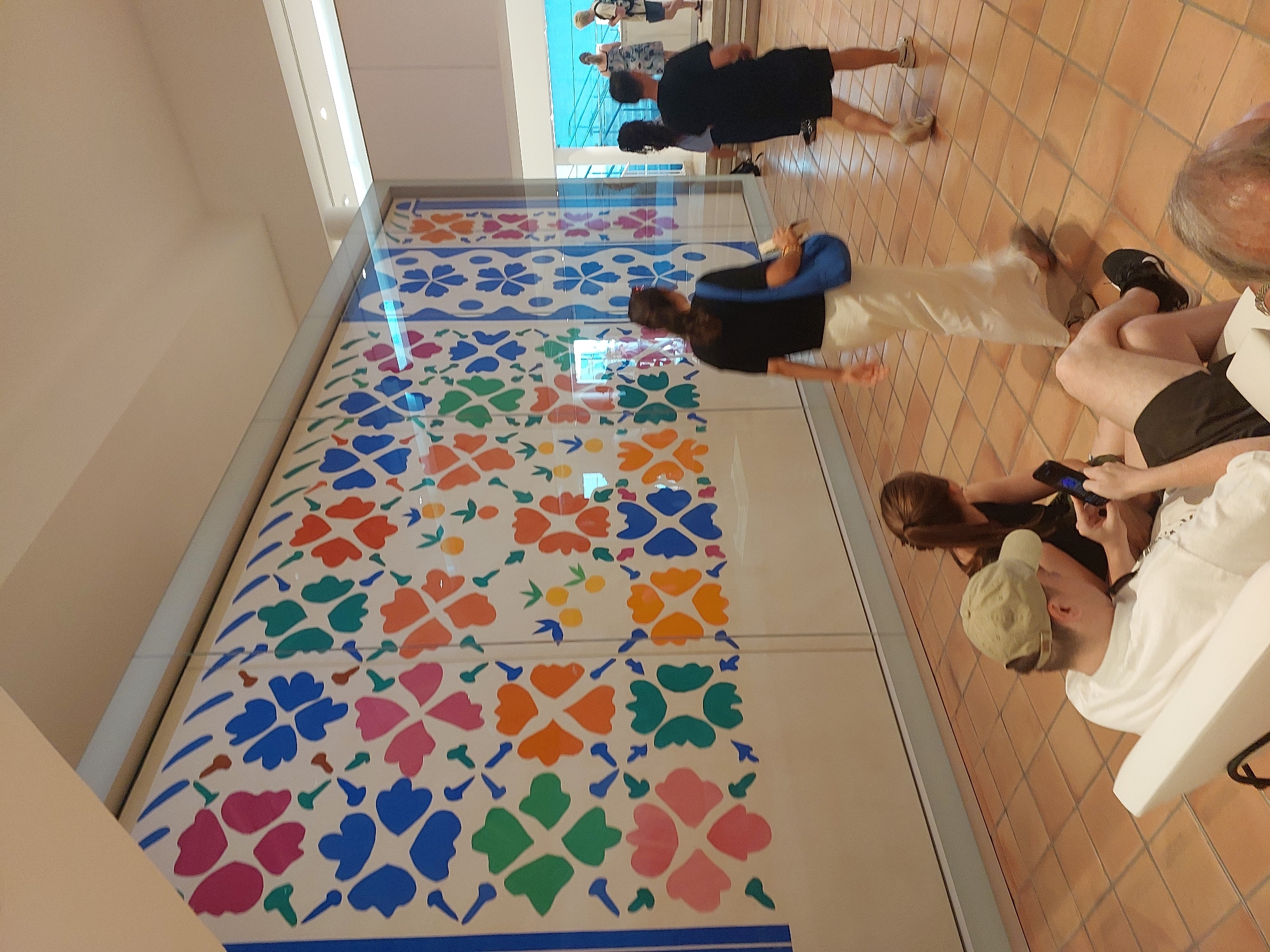 In Nice, France, inside a brightly lit exhibition space, a large, colorful mural adorns a wall. The mural features a repeating pattern of stylized flowers and fruit in vibrant shades of blue, pink, orange, and green against a white background, bordered by vertical blue stripes on the left. In the foreground, two people are seated on the floor, one a male wearing a light-colored t-shirt and a khaki baseball cap, holding a black smartphone. Beside him, a female is seated, looking down at her phone. A white bench is partially visible. Moving further into the space, a woman in a black top and a long, flowing cream-colored skirt walks away from the viewer, carrying a bright blue bag. To her right, a man in a black t-shirt and black shorts walks towards the viewer, also wearing athletic shoes. In the background, other individuals are visible near a staircase leading to a blue-tiled area. The floor is tiled with terracotta-colored squares.