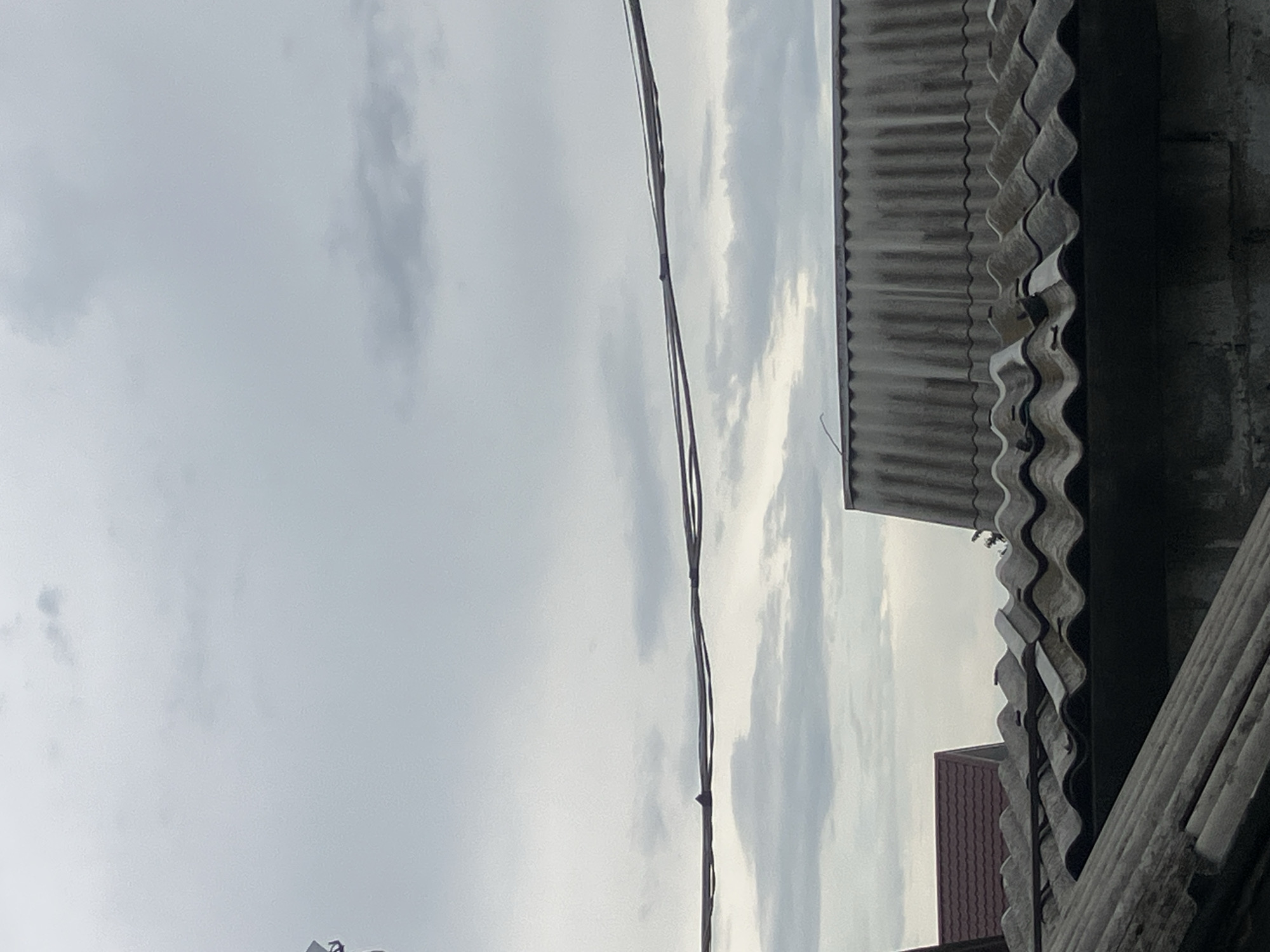 The image shows a cloudy sky above several rooftops in Vovkivka, Ukraine. Two prominent electrical wires run horizontally across the frame. Below the wires, a corrugated metal roof with a visible ripple pattern is seen, extending across the lower half of the image. To the left, a section of a darker-colored, possibly brick, wall is visible. Behind this, a roof with red, textured shingles is partially visible. Further to the right and behind the central corrugated roof, another corrugated roof with a more aged, weathered appearance is dominant. The overall scene depicts an outdoor, possibly residential or industrial, area under an overcast sky.