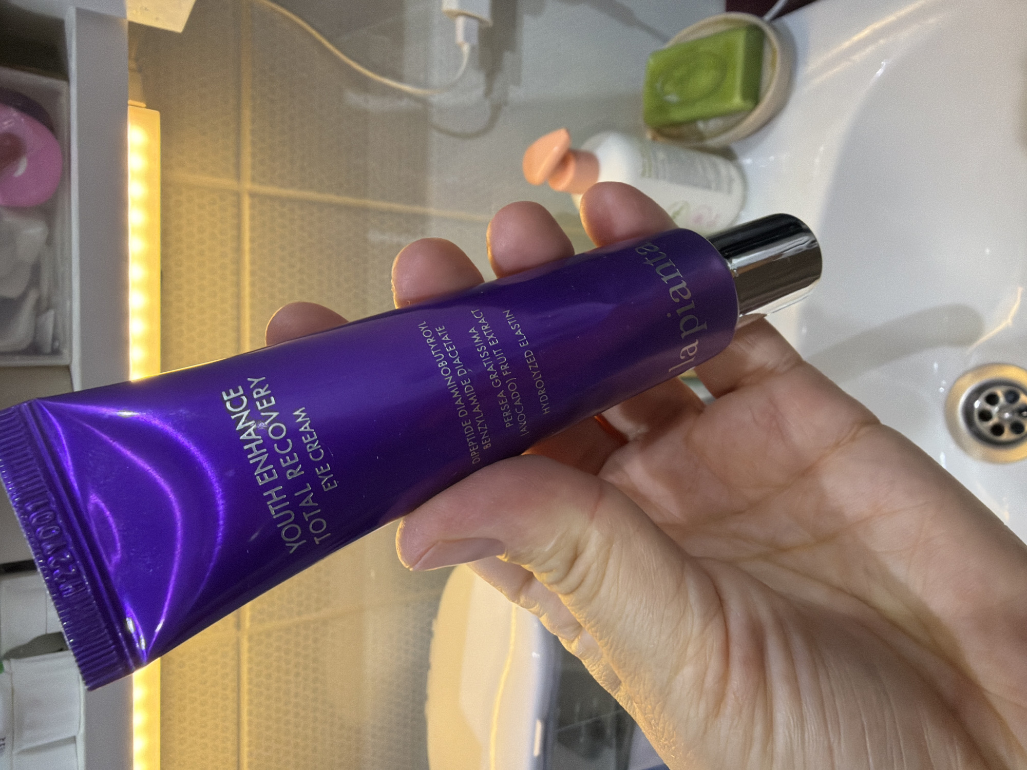 A person holds a purple tube of "la pianta" Youth Enhance Total Recovery Eye Cream in their hand. The tube is almost entirely in focus, while the background is somewhat blurred. The background features a bathroom sink with a green bar of soap and various other toiletries. The location is likely a private residence. There is no indication of the city of Chaikovsky, Russia.