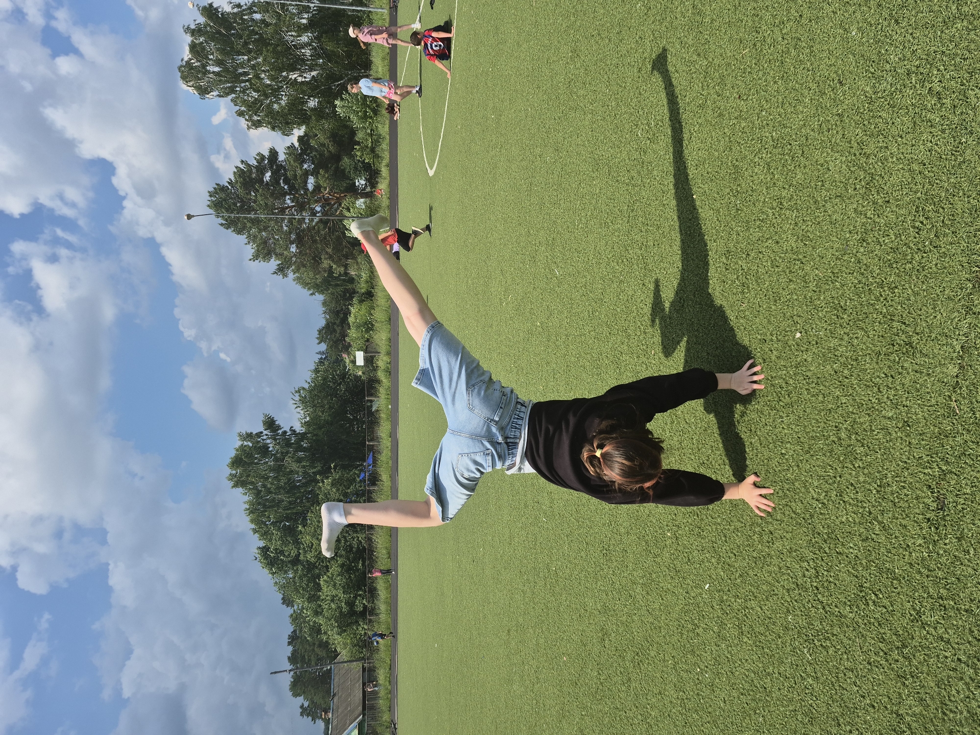 An outdoor, full-shot image on a sunny day shows a young person performing a handstand on an artificial turf field in Chaikovsky, Russia. The person, wearing a black long-sleeved shirt and denim shorts, is in mid-handstand with their legs extended upwards, one bent and the other straight. Their hands are on the ground, and their shadow is cast to the right. In the background, several other children are present on the field, some standing and others sitting or lying down. Trees and a building are visible in the distance under a partly cloudy sky.