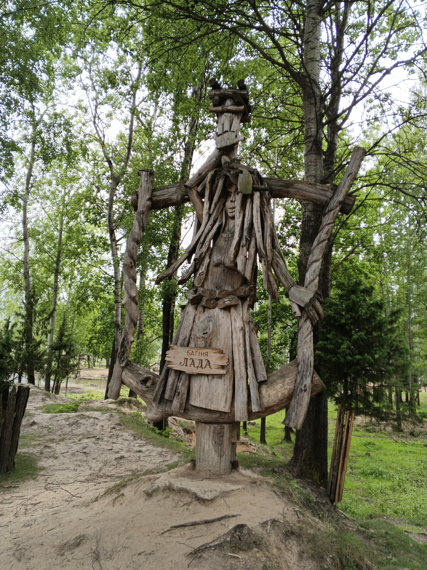 The central subject of the image is a large, intricately crafted wooden sculpture located outdoors in a lush, wooded environment. This towering figure, constructed from various logs, branches, and planks, depicts what appears to be a humanoid deity. Its head is composed of stacked wooden pieces, with a carved, somewhat serene face visible on its chest, covered by hanging wooden strips resembling hair or garments. The main body is formed from vertical wooden planks, giving it a robe-like appearance, while thick, twisted logs serve as its outstretched, bent arms. The sculpture stands on a base of logs atop a small sandy mound, next to a dirt path.
A small wooden plaque is affixed to the sculpture's lower torso, bearing text in Cyrillic. The inscription reads "БАГІНЯ ЛАДА," which translates to "Goddess Lada," identifying the figure as the Slavic deity of harmony, beauty, and fertility.
The surrounding scene is characterized by dense foliage. Tall deciduous trees with vibrant green leaves dominate the background and frame the sculpture, their canopies allowing bright daylight to filter through. In the distance, beyond the path and grassy areas, a body of water, possibly a river or lake, is visible, bordered by more trees. The bright, clear lighting suggests it is daytime, likely morning or afternoon, with pleasant weather. The setting appears tranquil and natural, located in Малахавецкі сельскі Савет, Belarus.