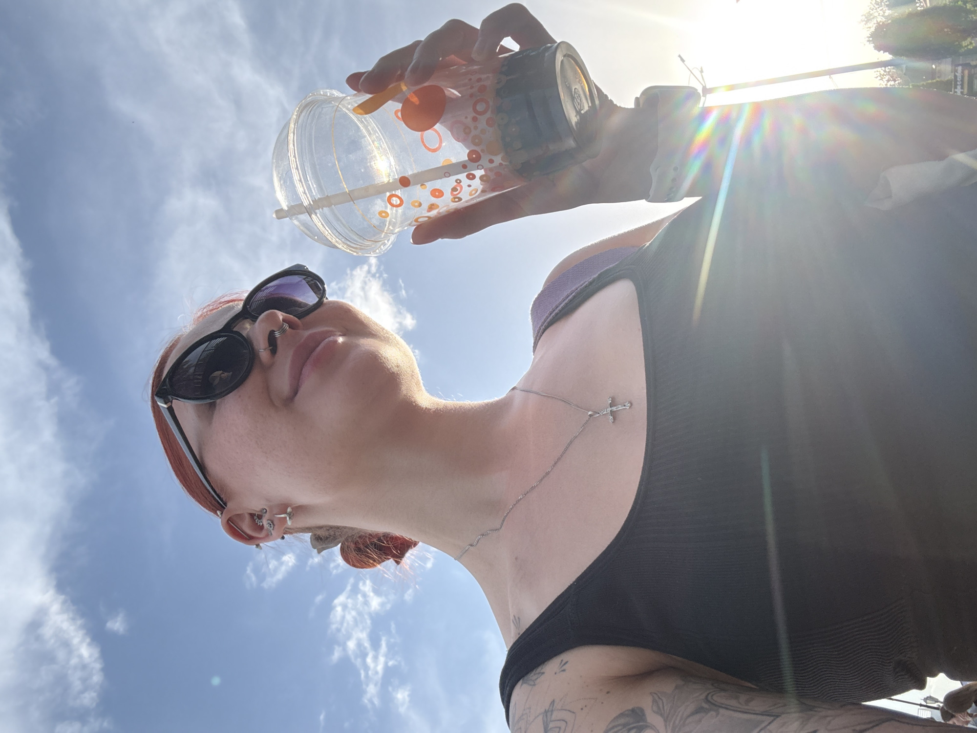 A low-angle shot depicts a person with fair skin, wearing dark sunglasses and a black tank top, holding a plastic cup with a lid and straw against a bright, sunlit sky with scattered clouds. The person has red hair, multiple piercings in their ear, and a silver necklace with a cross pendant. Tattoos are visible on their arm. The location is Rostov-on-Don, Russia.