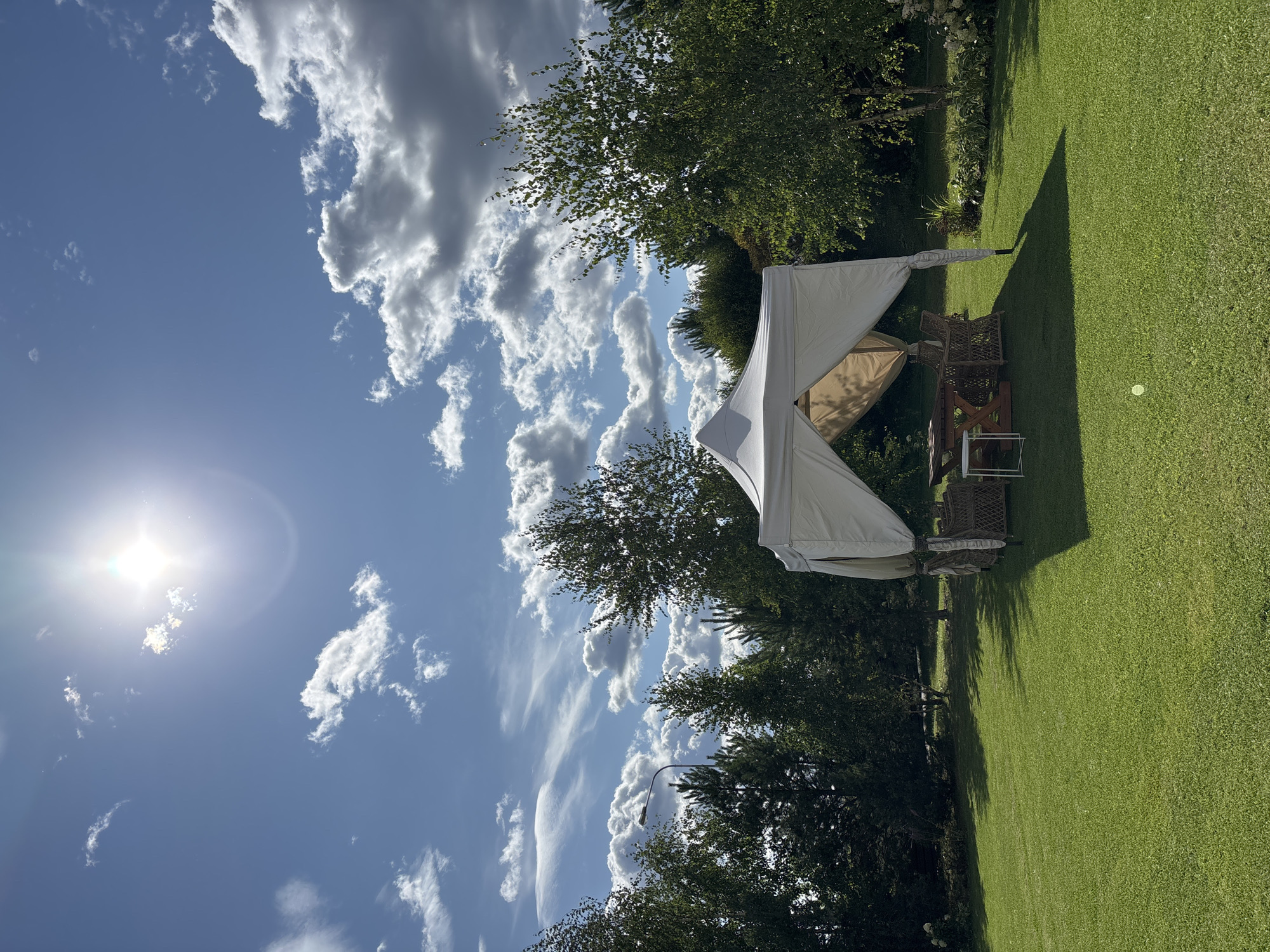 This is an exterior, eye-level, wide shot of a grassy backyard in Pokrovskoe, Russia, on a sunny day with some clouds. In the center of the frame is a white canopy tent with wicker furniture, a wooden table, and a white stool inside. Surrounding the tent are trees, with a lamp post visible in the background. The sun is shining brightly in the upper portion of the image, with scattered clouds. The grass is well-maintained.