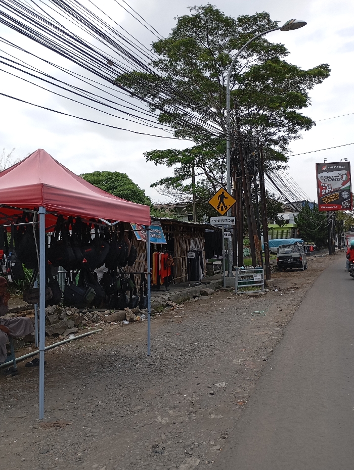The image shows a street scene in Katapang, Indonesia (latitude: -6.99, longitude: 107.557). A street vendor is selling backpacks from a red tent canopy in front of a building. The vendor has several bags on display and a person is sitting in the tent near the bags. A pedestrian crosswalk sign stands in the middle of the street with a yellow background and a black silhouette of a person walking. A large tree with several branches is in the center of the image. The street is lined with utility poles with many wires running overhead. In the distance, a car and a motorcycle are traveling in the opposite direction. There is a building with a small sign near the street. The building has a bamboo fence and a few clothes are hanging on it. The ground surface is mostly gravel with some areas of pavement. The sky is overcast with a few clouds.