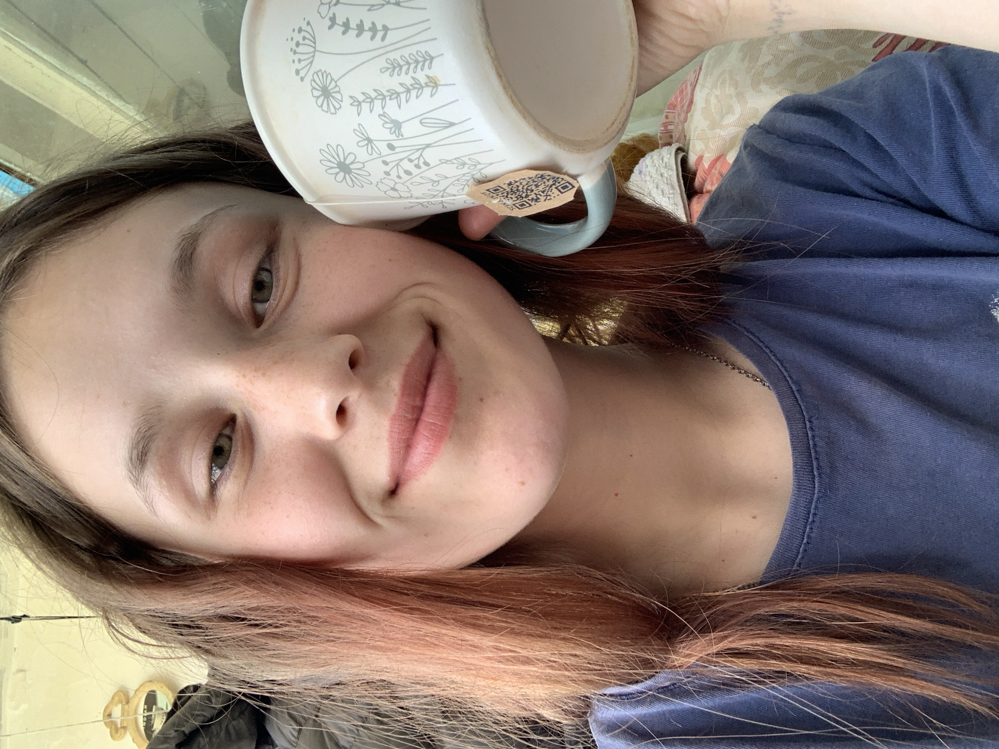 A young woman with light skin, brown hair with reddish highlights, and hazel eyes smiles while holding a white mug with a floral pattern and a light blue handle against her head. The mug has a tag with a QR code attached to the handle. She is wearing a blue t-shirt and a silver necklace. The background includes a patterned pillow, a window, and a glimpse of a yellow wall with a decorative item. There is no indication the image was taken in the city of Фоки, Russia.