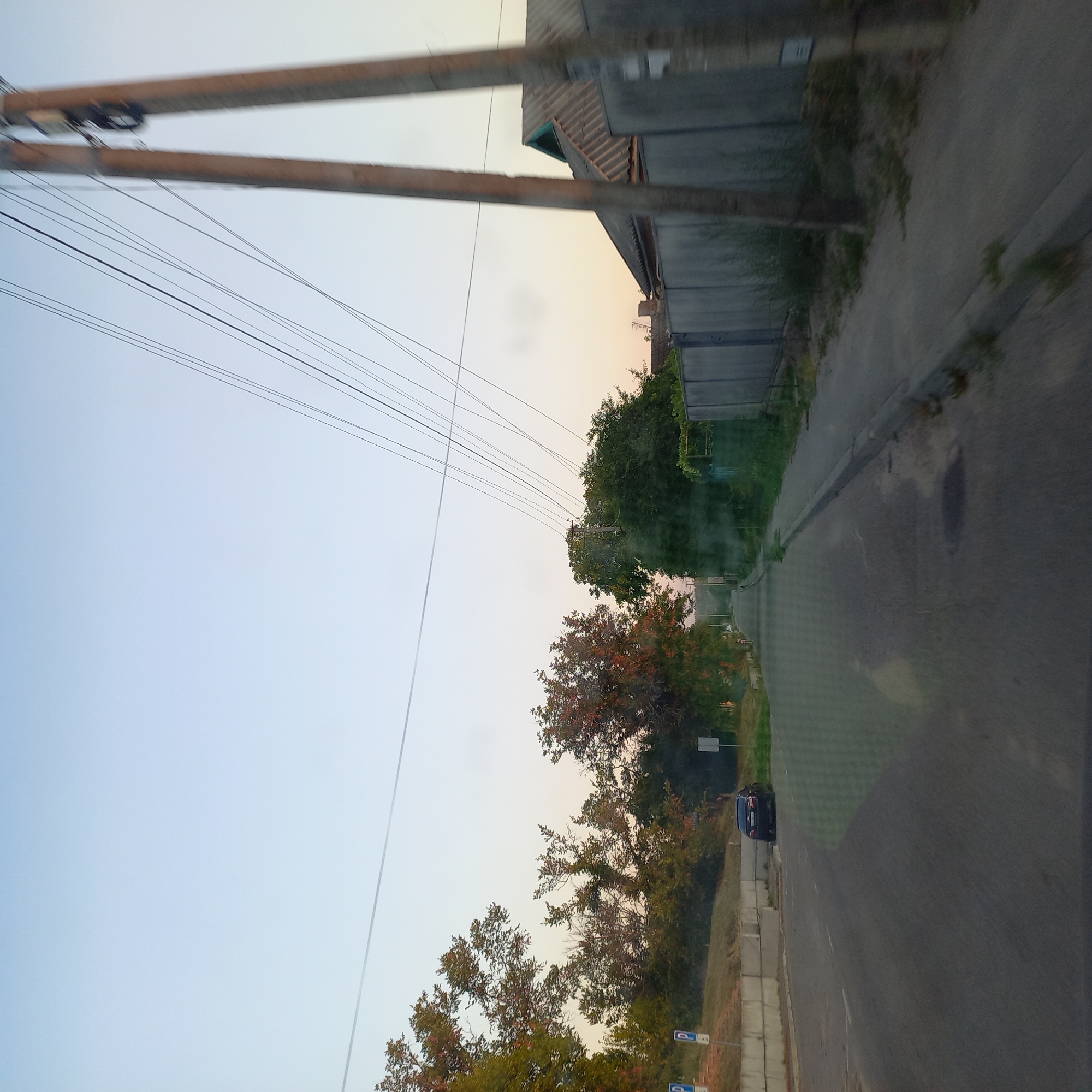 The image shows a street scene in Soniachne, Ukraine. The road is paved and appears to be in a residential area. There are trees on both sides of the street, with some showing autumn colors. Power lines are visible overhead, supported by wooden poles. A gray fence runs along the right side of the street, with a house visible behind it. A black car is parked on the side of the road on the left. The sky is clear and light blue, suggesting it is either early morning or late afternoon. There are no people clearly visible in the immediate foreground. The scene depicts a quiet street in a Ukrainian town during the fall season.