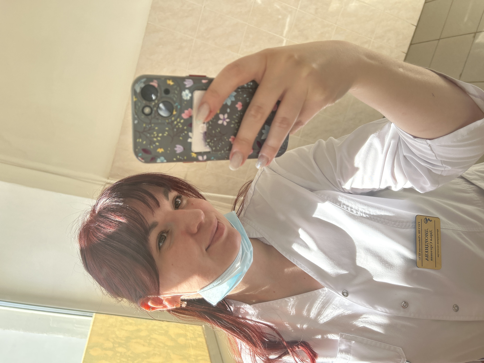 A woman in a white medical coat is taking a selfie indoors. She is wearing a light blue surgical mask pulled down below her mouth. She has red hair with bangs and is holding a phone with a floral case. A name tag is visible on her coat, partially obscured, but the name "Yalovi S.D." and "LD3 (IK 'Dubrovskaya T.P.')" can be seen. The background appears to be a hallway with tiled walls. The location is Фоки, Russia.