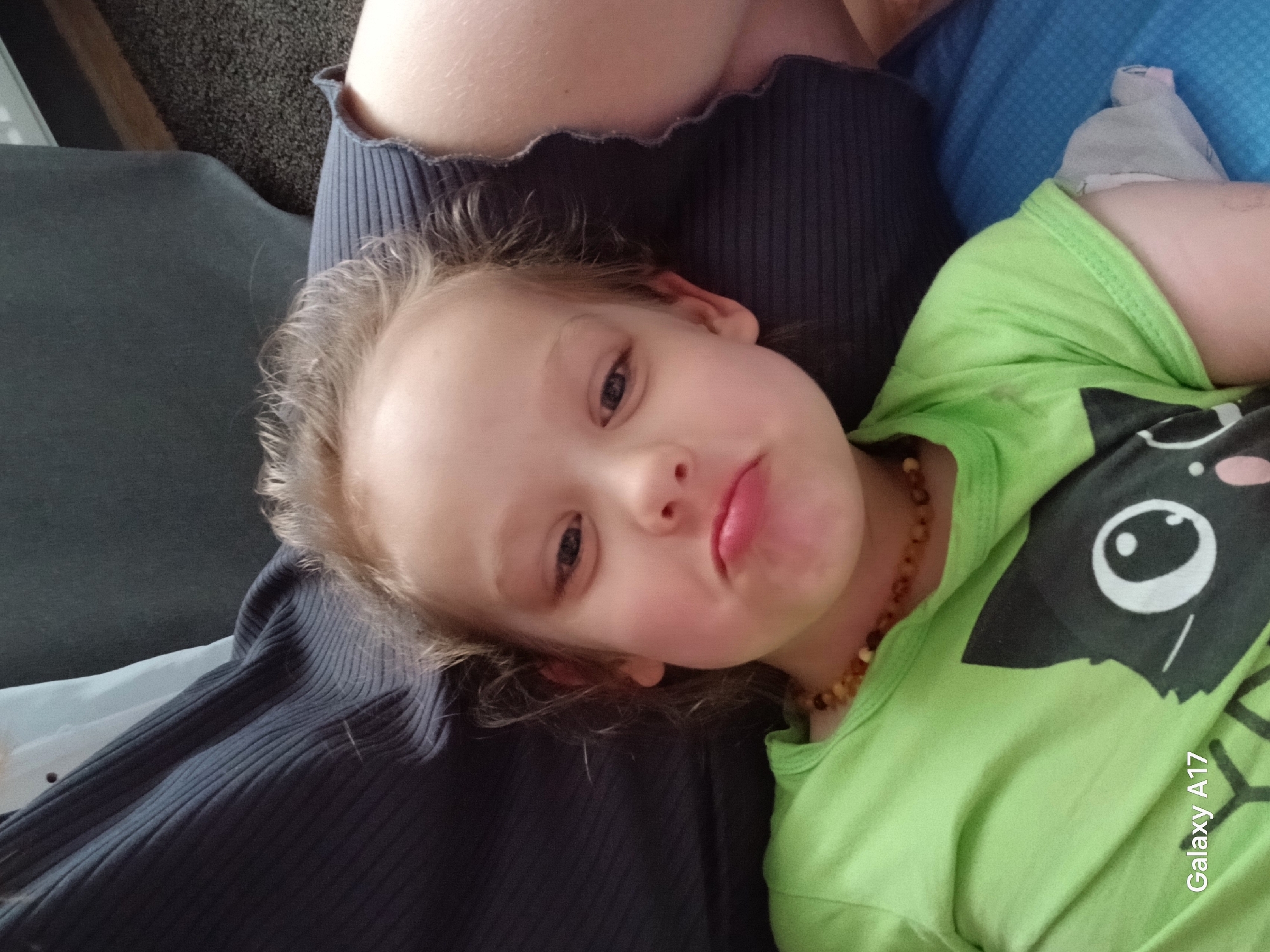 A young child is resting on someone's lap. The child has fair skin, light blonde hair, and is wearing a bright green t-shirt with a black and white graphic. The child's face is turned towards the camera with a slight pout. The adult's clothing appears to be a dark, ribbed garment. A light blue blanket or sheet is visible in the background. The "Galaxy A17" watermark is visible in the lower right corner of the image. The location in Yekaterinburg, Russia cannot be determined from this image.