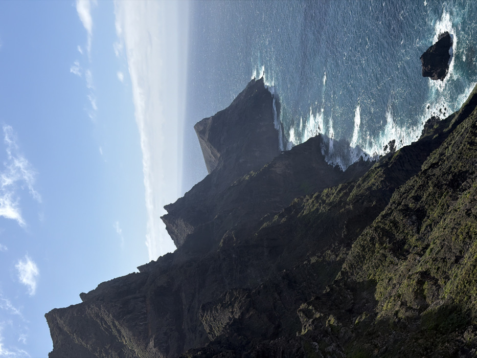 The image displays a rugged, unpopulated coastal landscape, identified as Tirma, Spain. The left two-thirds of the frame are dominated by a series of steep, dark grey-brown volcanic cliffs that descend sharply into the ocean. These formidable cliffs feature jagged peaks, pronounced ridges, deep shadows, and visible stratification, with scattered patches of dark green, low-lying vegetation clinging to sun-exposed slopes. A narrow, lighter-colored horizontal geological indentation is visible on one of the mid-ground cliff faces. The upper third of the image shows a clear blue sky with sparse, scattered white cumulus clouds. The Atlantic Ocean occupies the lower right section, presenting a deep blue hue with turquoise undertones and active wave patterns. White foam from breaking waves is evident along the rocky shoreline, with whitecaps extending further into the open water. An isolated, dark rock formation protrudes from the ocean surface in the lower right quadrant. No human figures, animals, or artificial structures are present.