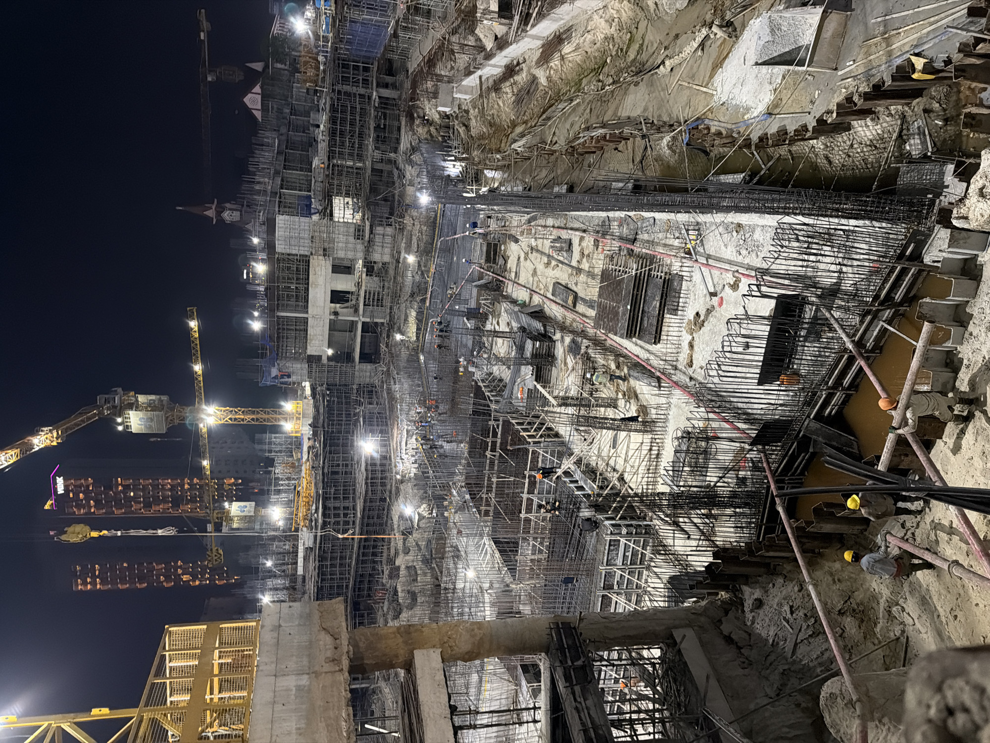 The image depicts a large construction site in Đà Nẵng, Vietnam, at night. There are several multi-story buildings under construction, heavily scaffolded with metal frameworks. Cranes are visible, illuminated by floodlights, alongside buildings and infrastructure. In the foreground, a deep excavation reveals soil and construction materials, with visible supports and reinforcements. Construction workers are present, wearing hard hats and working in the excavation. The image shows an active construction site with intense activity.