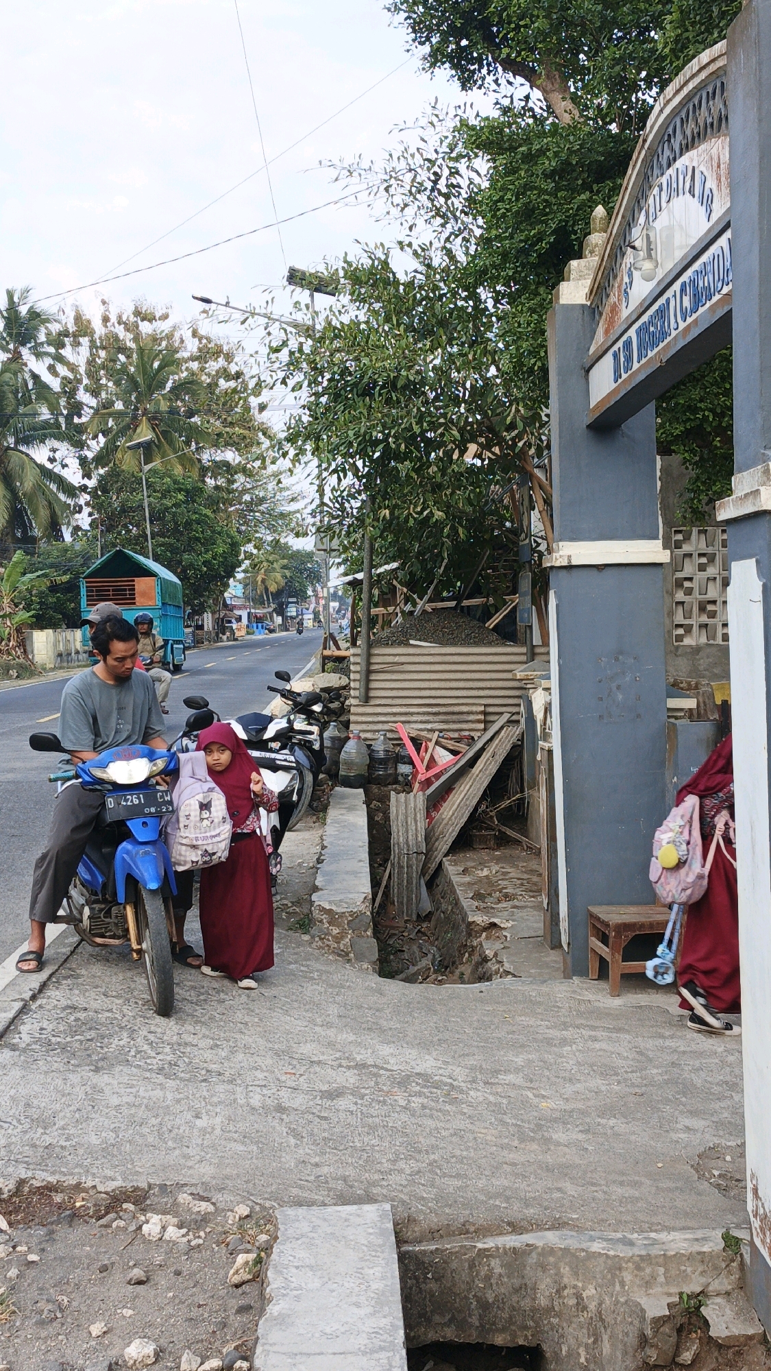 The image depicts an outdoor scene on a cloudy day in Cibenda, Indonesia. In the foreground, a man in a grey t-shirt and dark pants sits on a blue motorcycle, facing left. Beside him, a young girl wearing a burgundy hijab and dress, with a backpack on her back, stands facing the motorcycle. The motorcycle has the license plate "D 4261 CW 08-23". Behind them, another motorcycle is parked. To the right, a grey concrete archway with the inscription "SD NEGERI 1 CIBENDA" is visible. A portion of a building is also seen. The ground is paved with concrete, with some areas showing disrepair and dirt. To the left of the road, there are palm trees and other greenery. A blue truck is seen in the distance on the road. The overall environment suggests a street in a semi-urban or rural area, likely near a school.