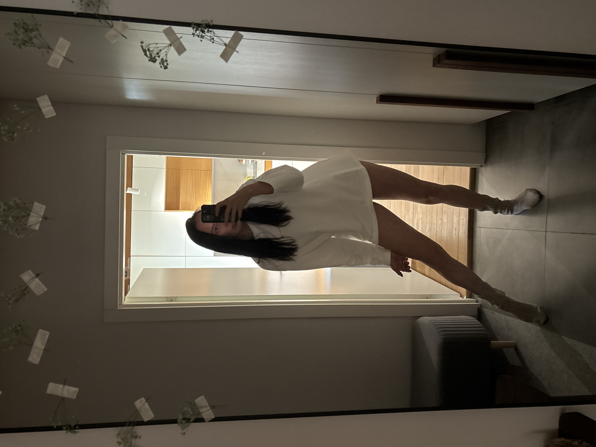 A person is taking a selfie in a mirror. The person is wearing a white oversized shirt and knee-high socks. The reflection shows a portion of a room. The room appears to have a doorway, a cabinet, a couch, and a tiled floor. The location is not discernible from the image.