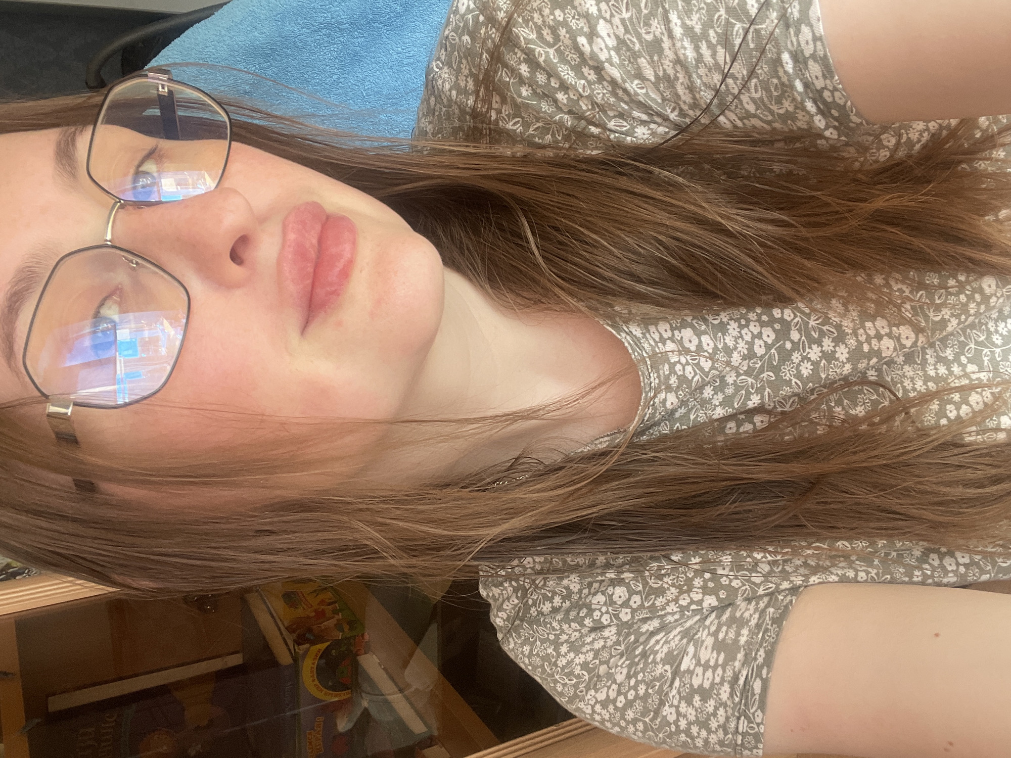 A young woman with long, light brown hair and glasses is captured in a close-up, chest-up selfie. She is wearing a light green t-shirt with a white floral pattern. Her hair is wet and appears to be styled down. The background shows a wooden bookshelf filled with books and a blue towel draped over a chair. The lighting suggests an indoor setting, possibly a home. The image does not contain any explicit information about the specific location in the city of Сырково, Russia, beyond what can be inferred from the general household environment.