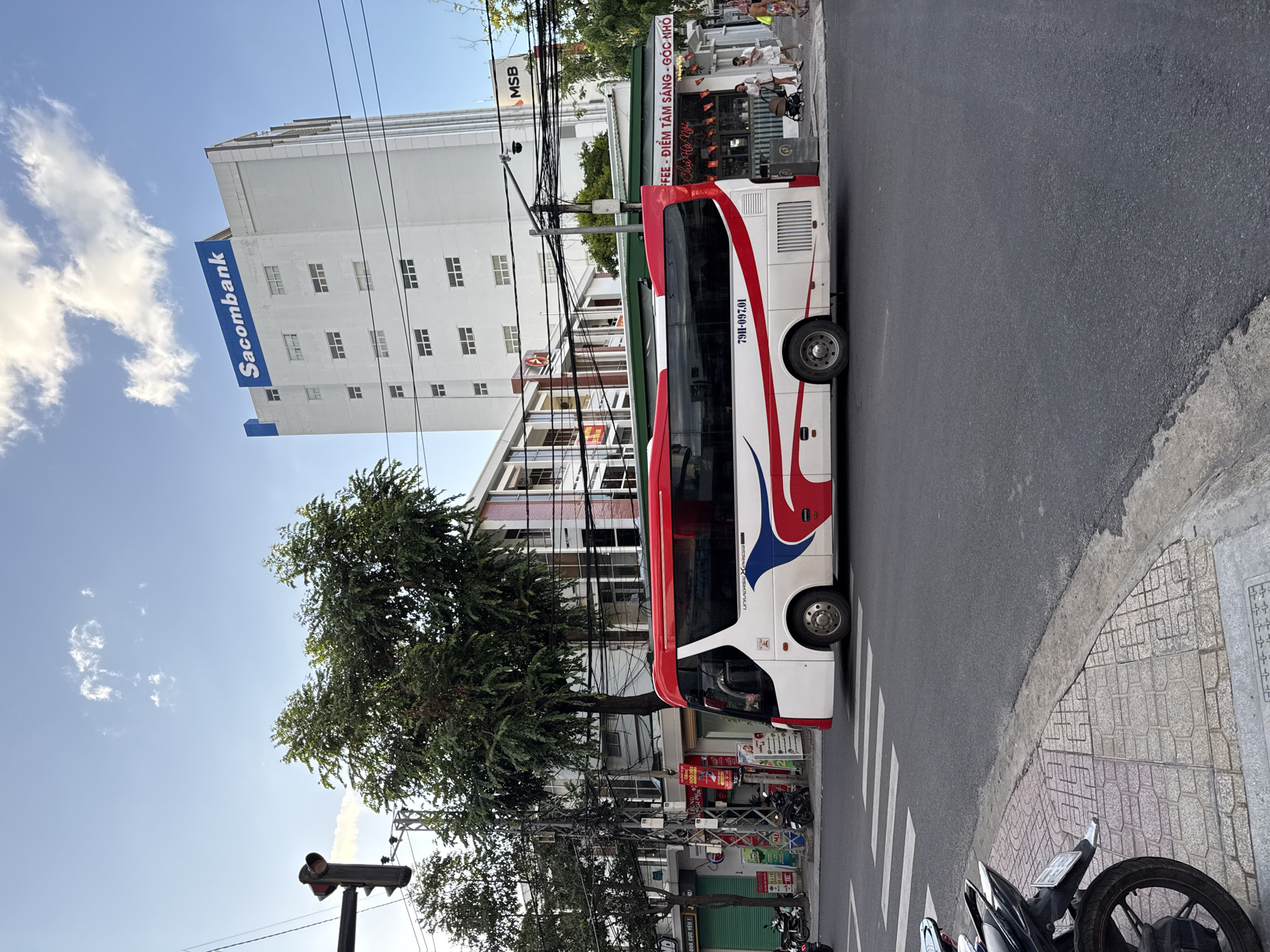 The image captures a street scene in Nha Trang, Vietnam, with a white and red bus prominently featured in the foreground. The bus, identified with the license plate "79H-097.01," is traveling on a paved road. Across the street, a multi-story building with the "Sacombank" logo is visible, along with other shops and businesses. Several trees line the sidewalk, and utility poles with numerous wires are present. A pedestrian crossing is marked on the road, and a motorcycle is parked on the sidewalk to the left. The sky is partly cloudy, with a bright, clear blue expanse. The overall atmosphere suggests a typical urban environment during the daytime.
