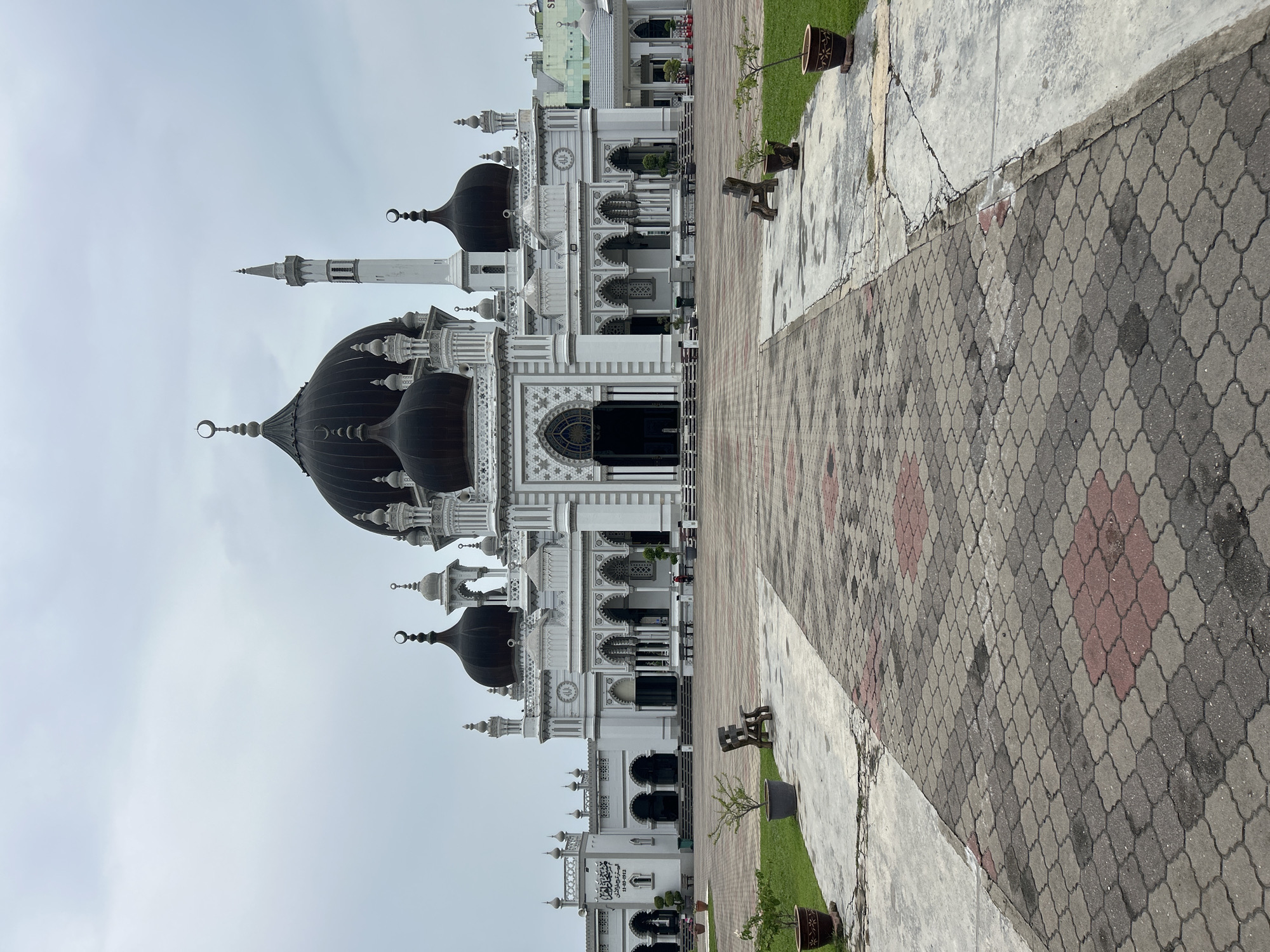 This is an exterior, ground-level, eye-level, full-frame, vertical shot of the Zahir Mosque in Alor Setar, Malaysia. The mosque is a prominent landmark, featuring a grand central dome and multiple smaller domes, all black. The mosque's facade is white with intricate grey detailing and arches, including a tall white minaret on the right side of the main structure. In front of the mosque is a large, paved courtyard. The paving stones are a mix of grey and red, arranged in a patterned walkway. Green grass areas are visible on either side of the walkway, with potted plants and wooden benches placed strategically. The sky is overcast with light grey clouds, suggesting a cloudy day. No people are clearly visible in the immediate foreground or performing any actions. The overall scene depicts the mosque and its surrounding plaza under a diffused light.