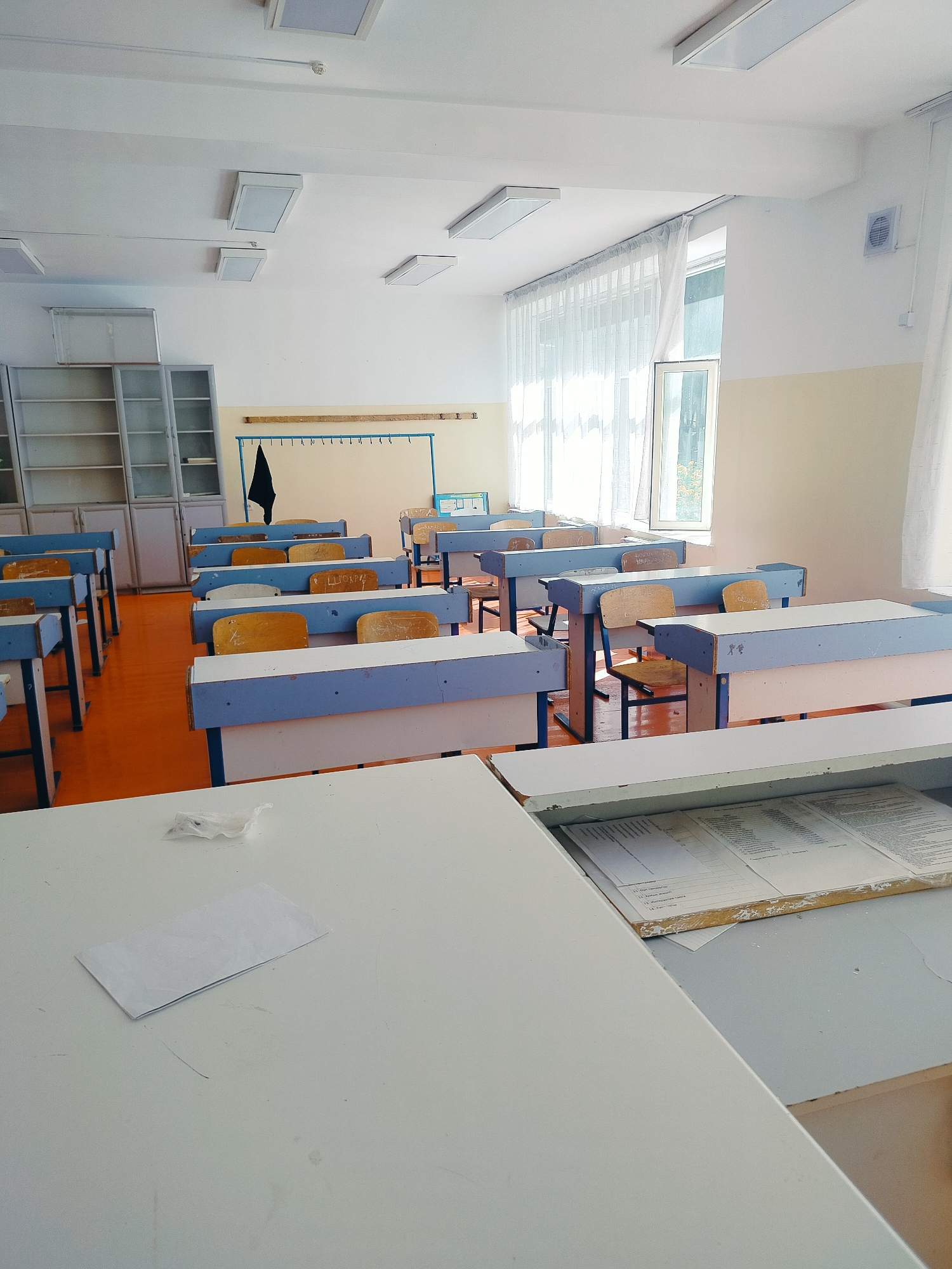 The image depicts an empty classroom in Көлкент, Kazakhstan. The room contains rows of desks with blue and white frames and light brown wooden seats and backrests. The floor is a vibrant orange. On the left side of the room, there are tall, grey metal bookshelves filled with various items. Along the back wall, there is a horizontal rack with hooks, and a black piece of fabric is hanging from it. A whiteboard is visible behind the desks. A large window with sheer white curtains is on the right side of the room, allowing natural light to enter. The ceiling is white and has several rectangular fluorescent light fixtures. In the foreground, there is a white desk with a folded piece of white paper and a crumpled white object on its surface. Another desk in the foreground has an open drawer containing documents.